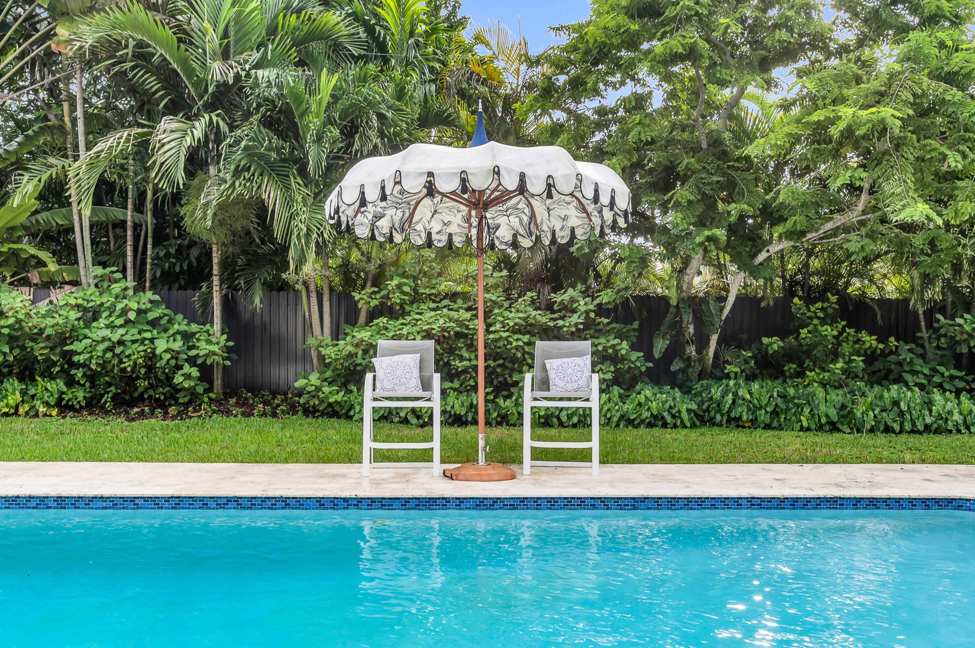 A white umbrella is positioned over two chairs beside a blue swimming pool, surrounded by lush greenery.