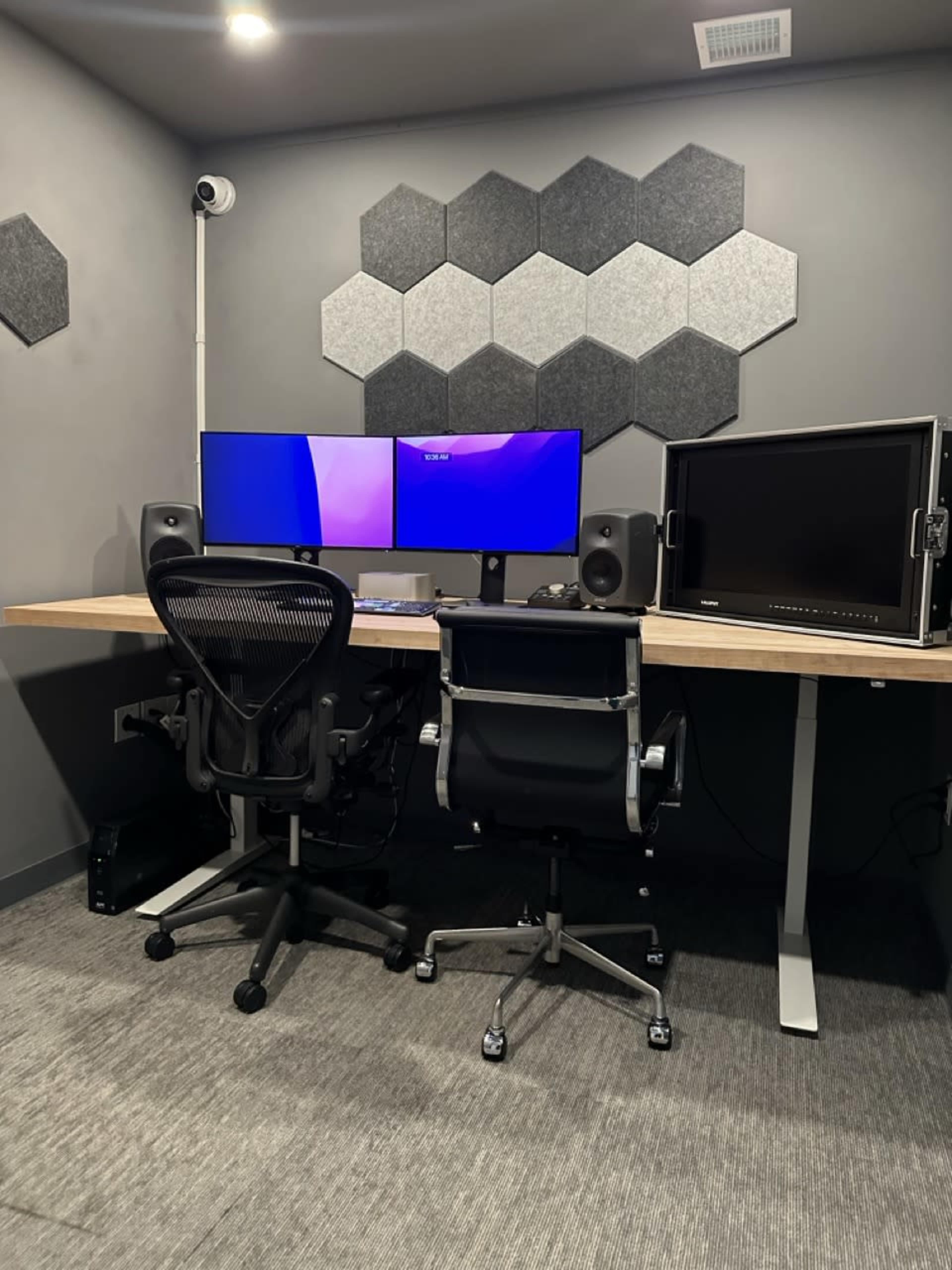 A modern workspace featuring a wooden desk with two computer monitors, speaker systems, and two ergonomic office chairs in a room with hexagon-shaped wall panels.
