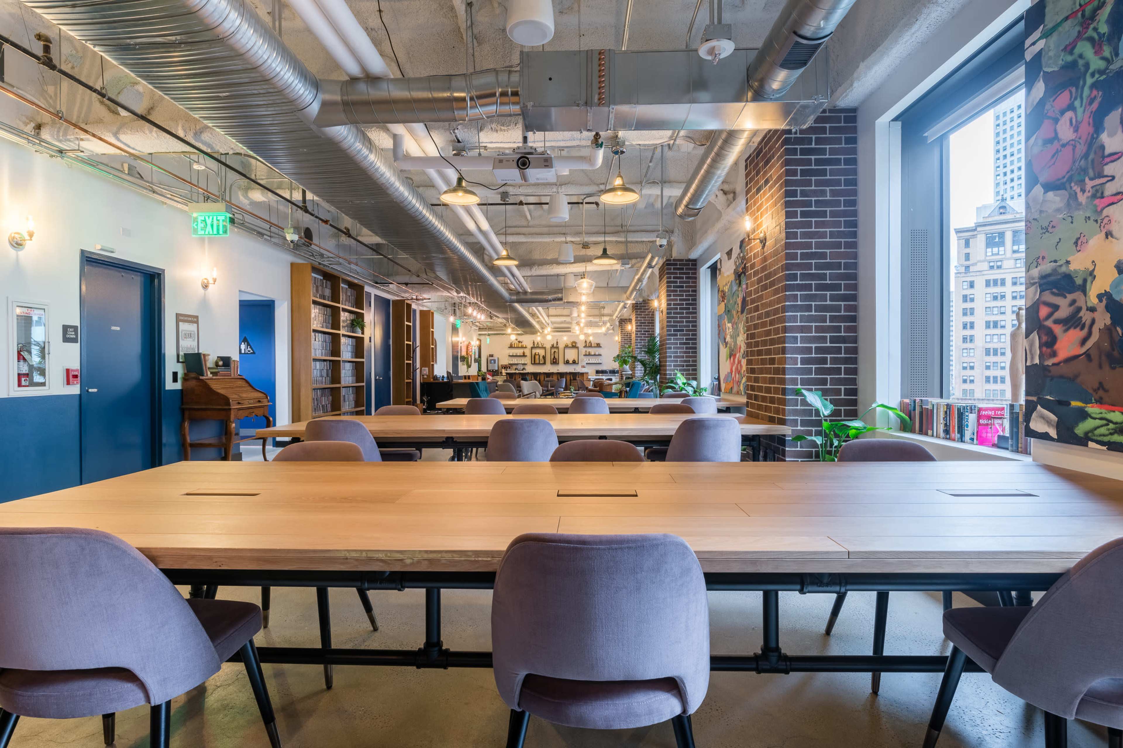 The image shows a spacious coworking area with large wooden tables, modern chairs, bookshelves, and large windows that let in natural light.