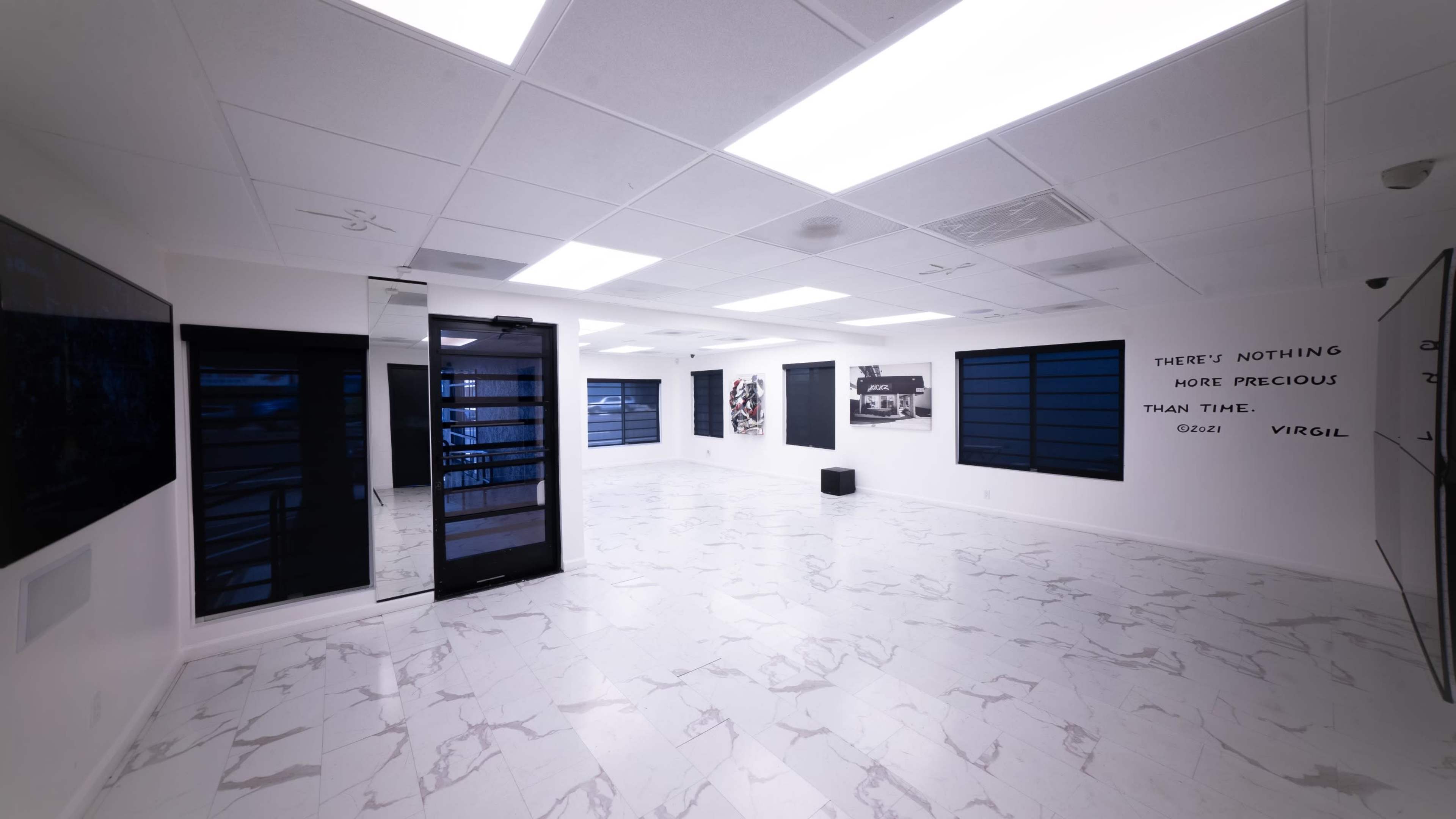 The image shows a spacious, well-lit room with white marble flooring, large windows, and minimalist decor featuring artworks on the walls.