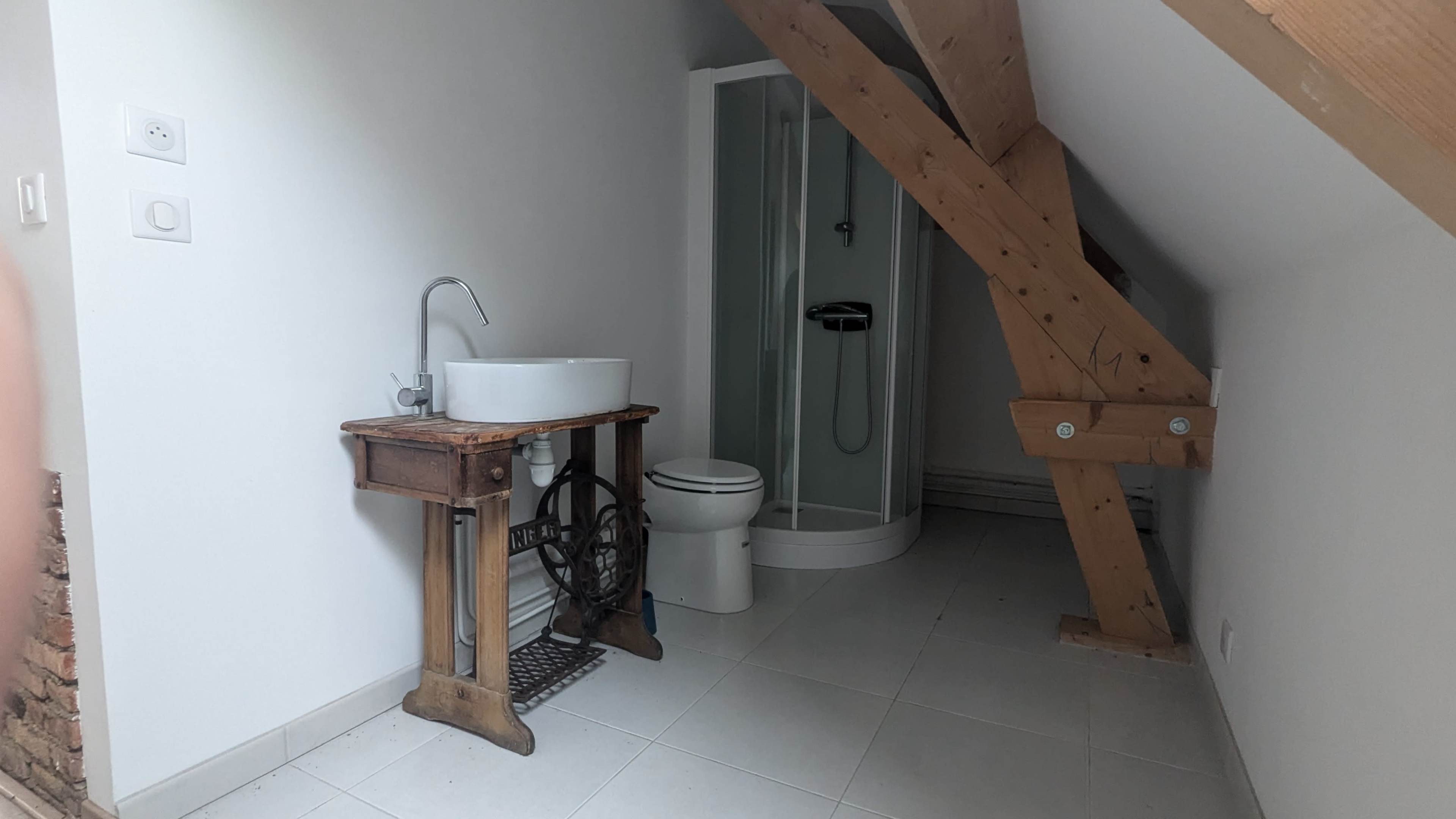 The image shows a small bathroom featuring a pedestal sink, a toilet, and a shower enclosed by glass, with wooden beams visible in the ceiling.