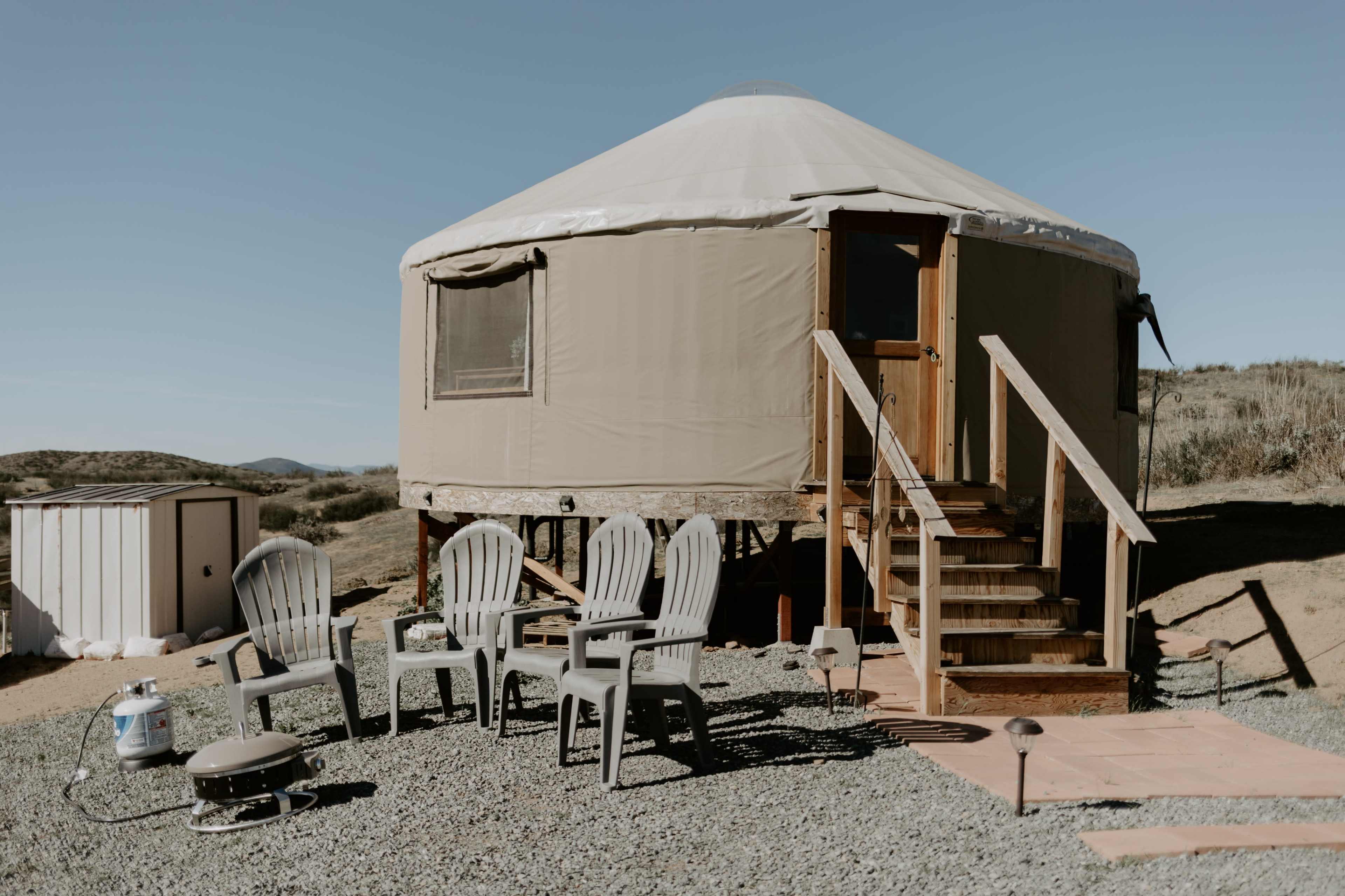 A yurt with a wooden deck and steps is surrounded by gravel and several plastic chairs.