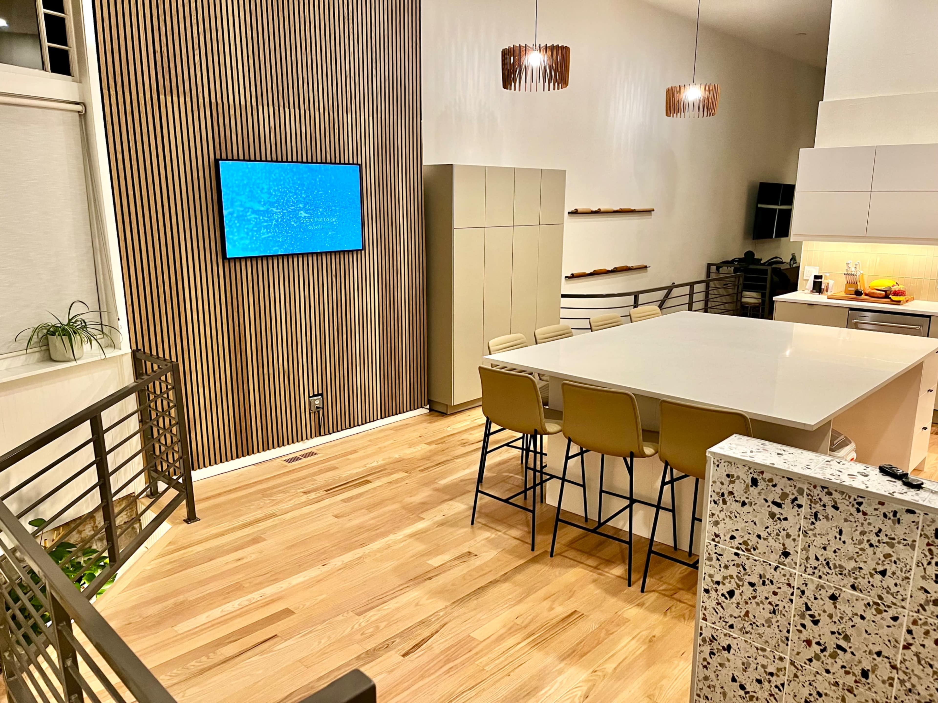 The image shows a modern kitchen with wooden flooring, a large island with bar stools, and a wall-mounted television displaying a blue screen.
