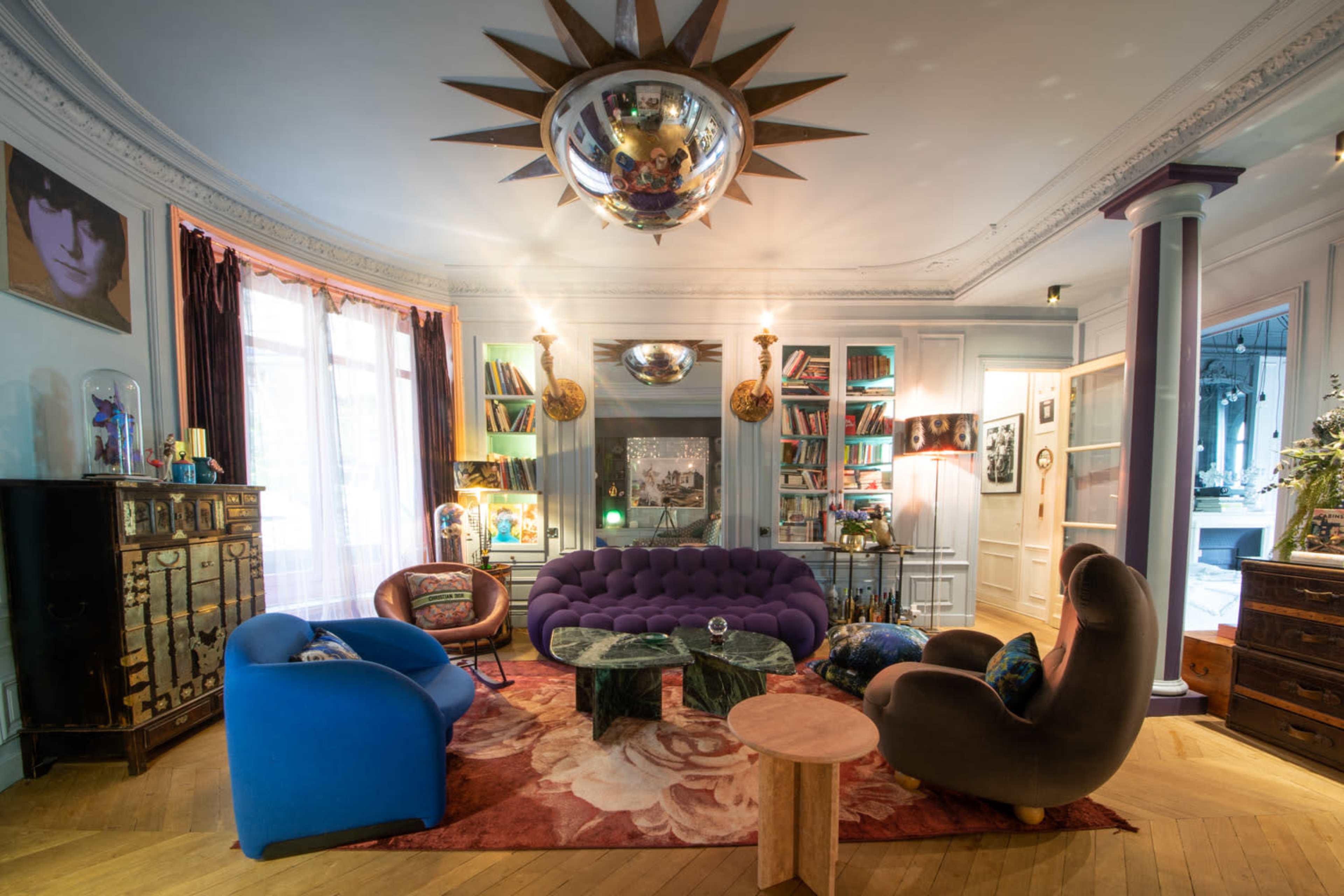 The image shows a stylish living room featuring a purple tufted sofa, two uniquely shaped armchairs, a round wooden coffee table, and a decorative ceiling fixture, all arranged around a floral area rug.