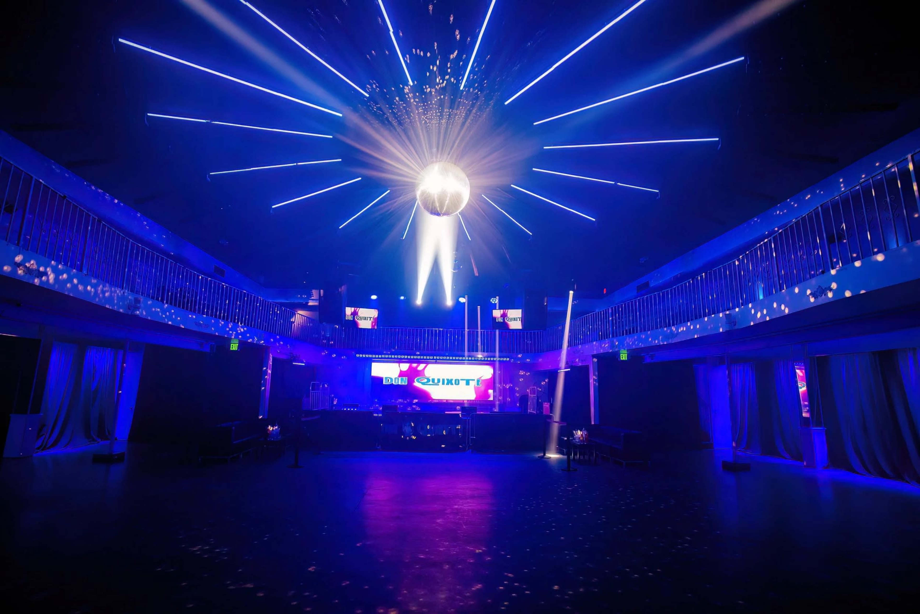 The image shows a dimly lit nightclub with a large disco ball hanging from the ceiling, surrounded by blue laser lights and a DJ setup in the background.