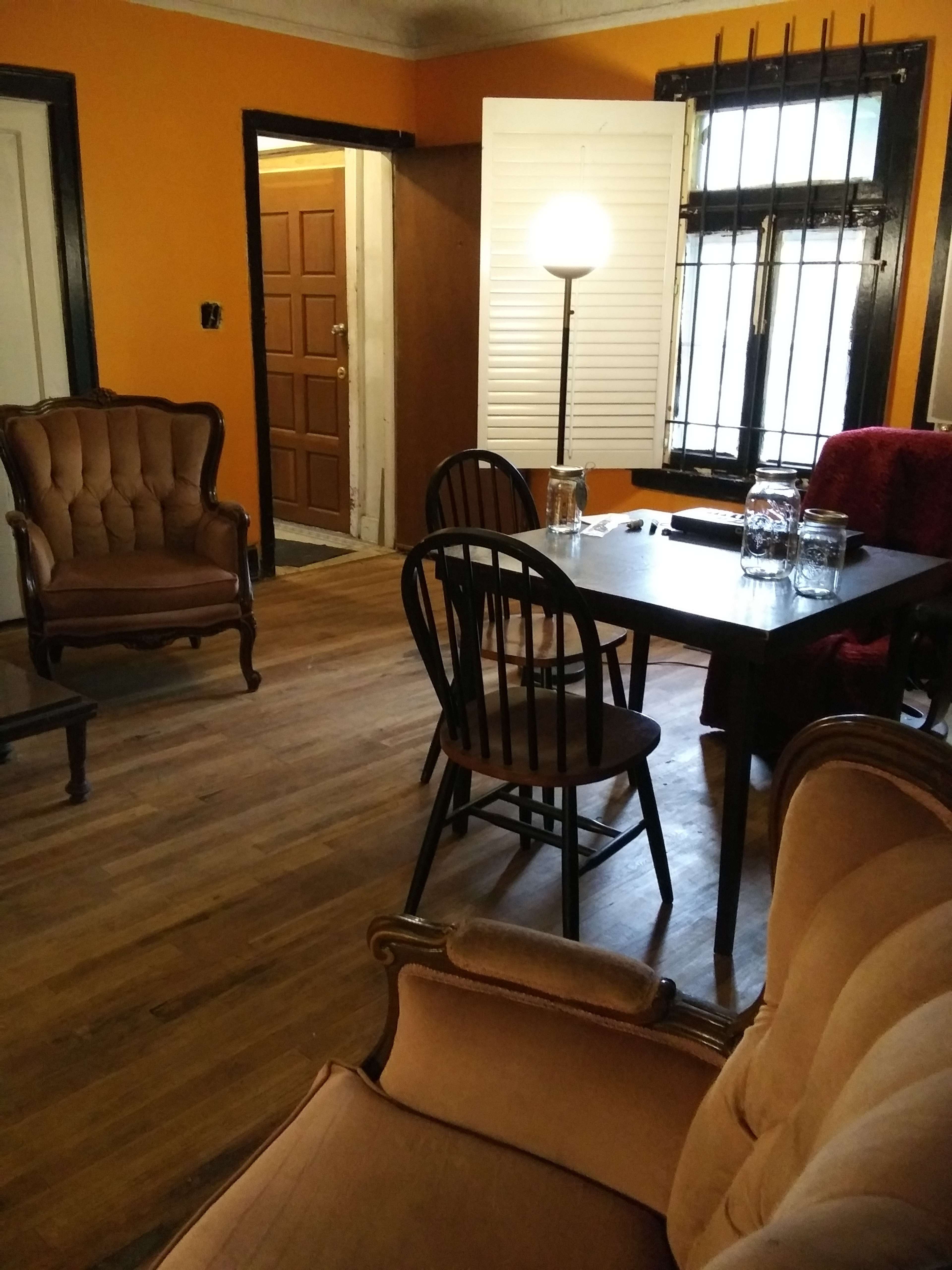 The image shows a living room with orange walls, a wooden floor, two upholstered chairs, a table with chairs, and a lamp in a corner near a window.