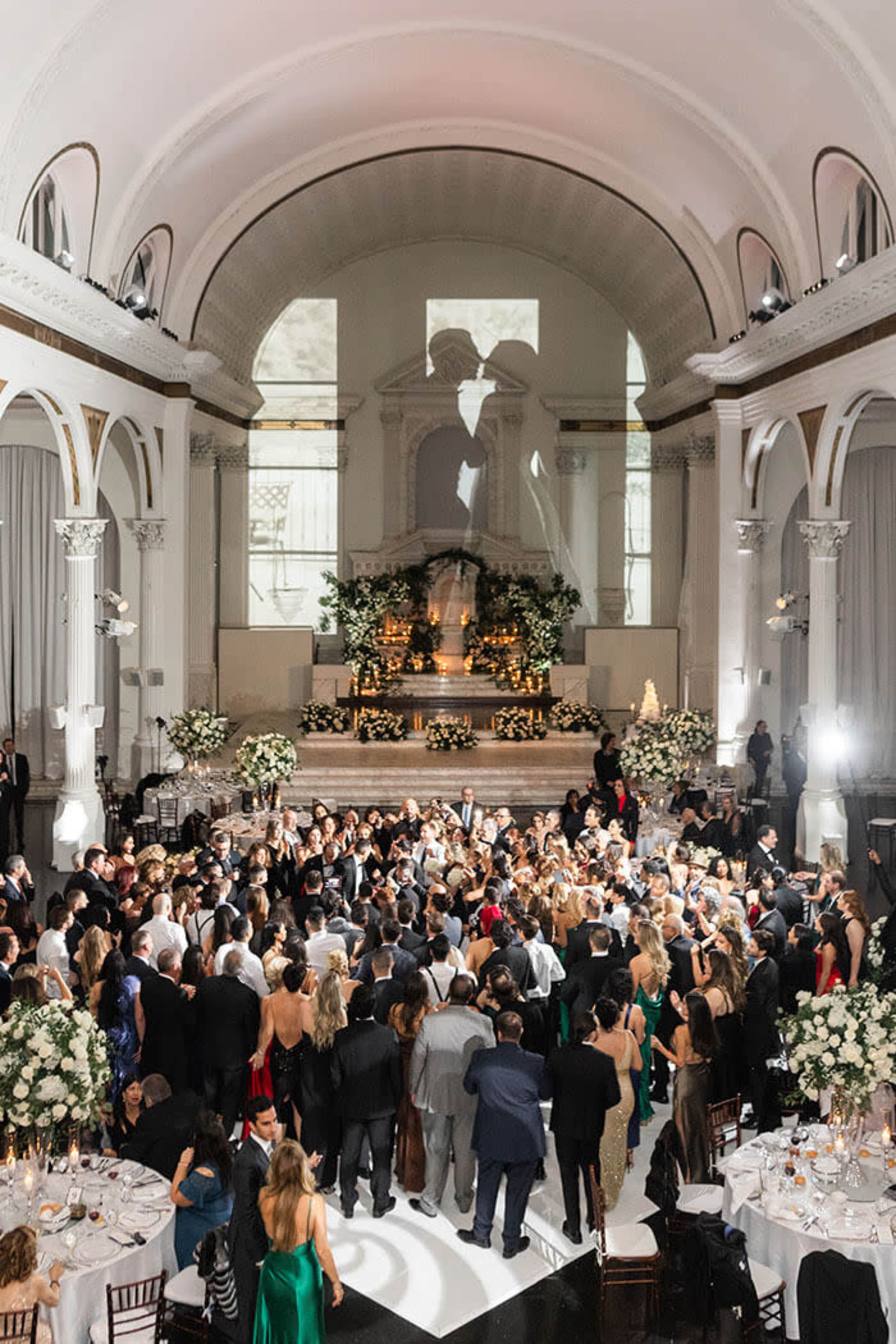 A large crowd gathers in an elegantly decorated venue with a projected silhouette of a couple on the wall.