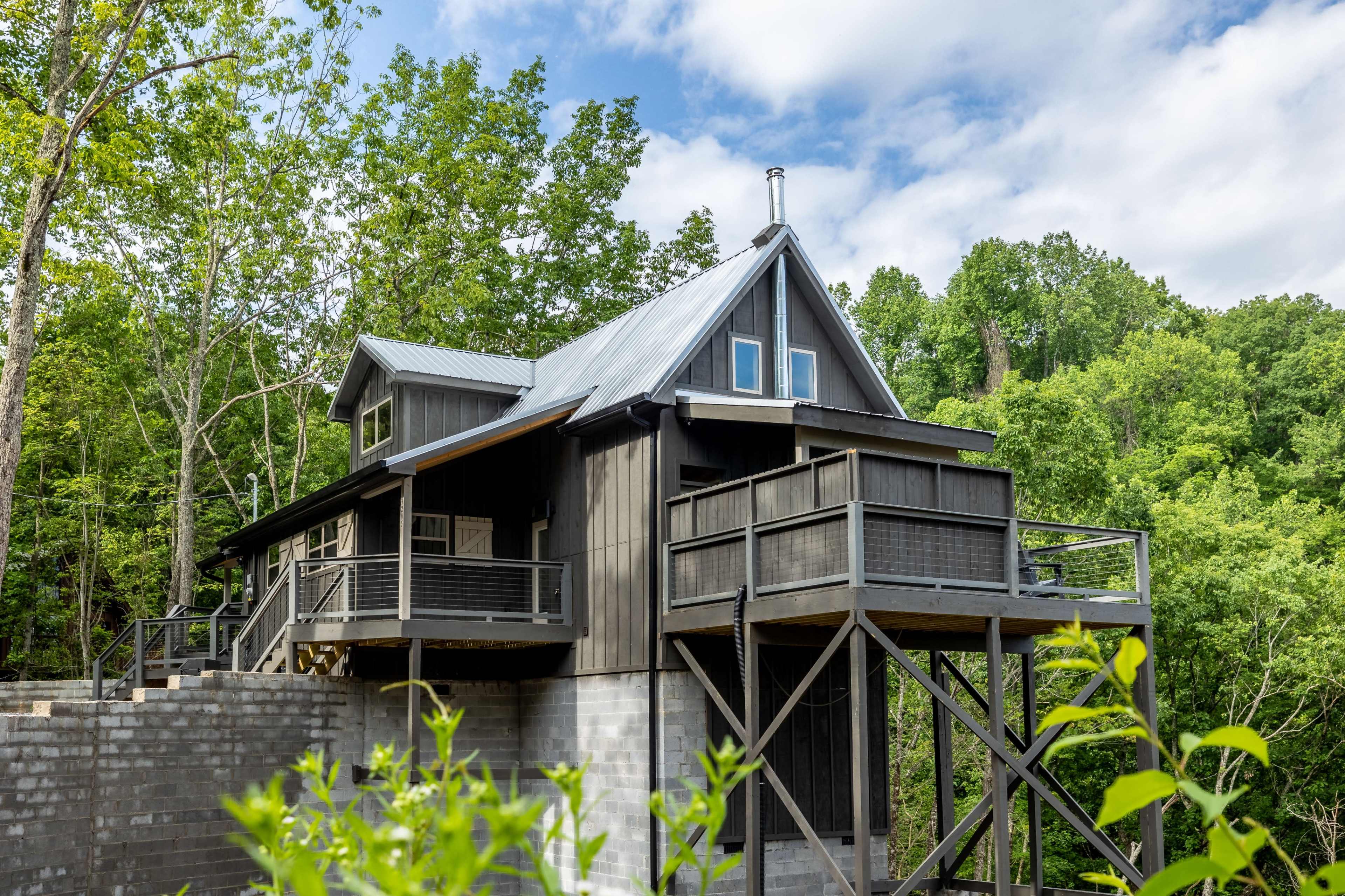 A contemporary house with a sloped roof and multiple balconies is situated on stilts among lush green trees.