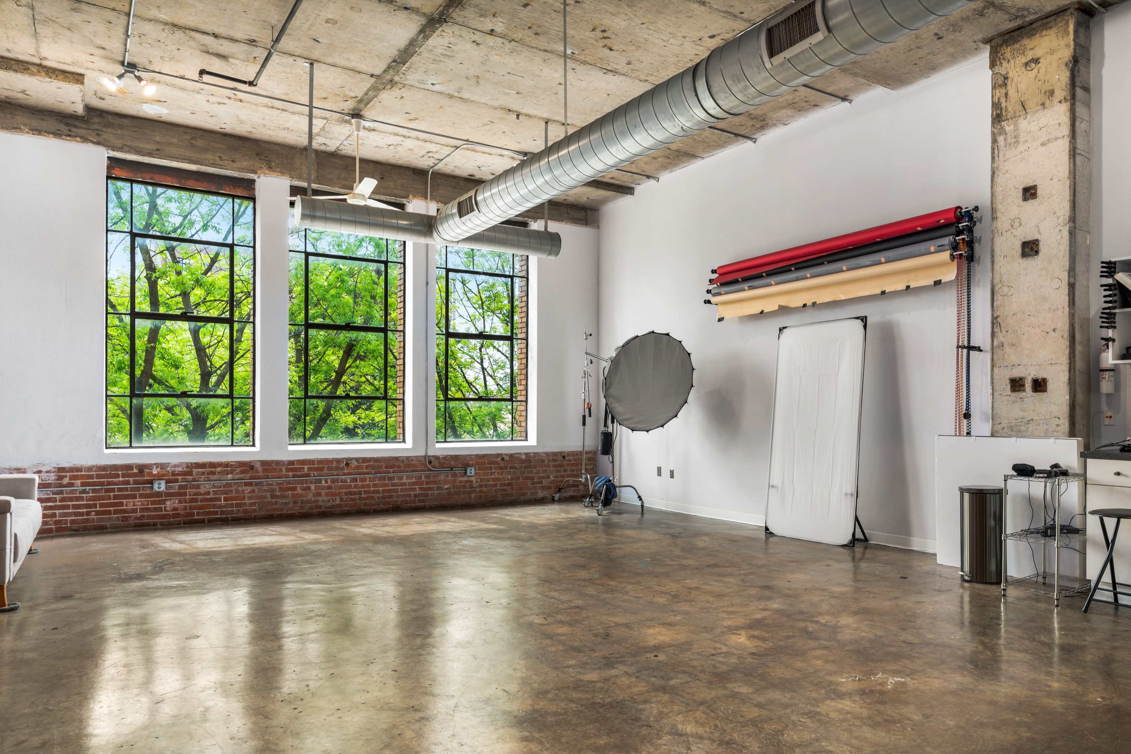 A spacious studio features large windows with views of green trees, exposed concrete ceilings, and photography equipment along the walls.