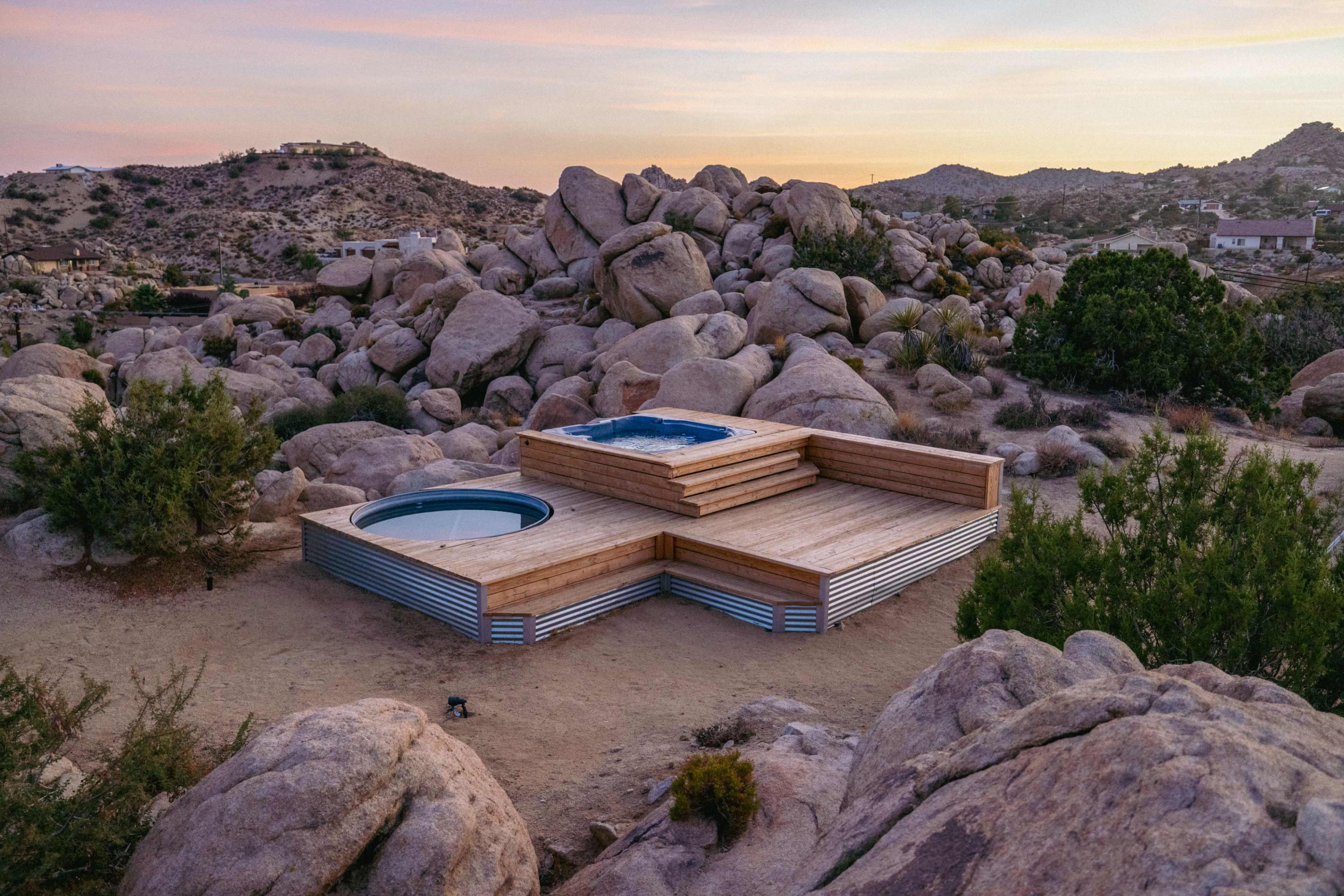 A wooden deck with a hot tub and a circular pool is nestled among large boulders in a desert landscape at sunset.