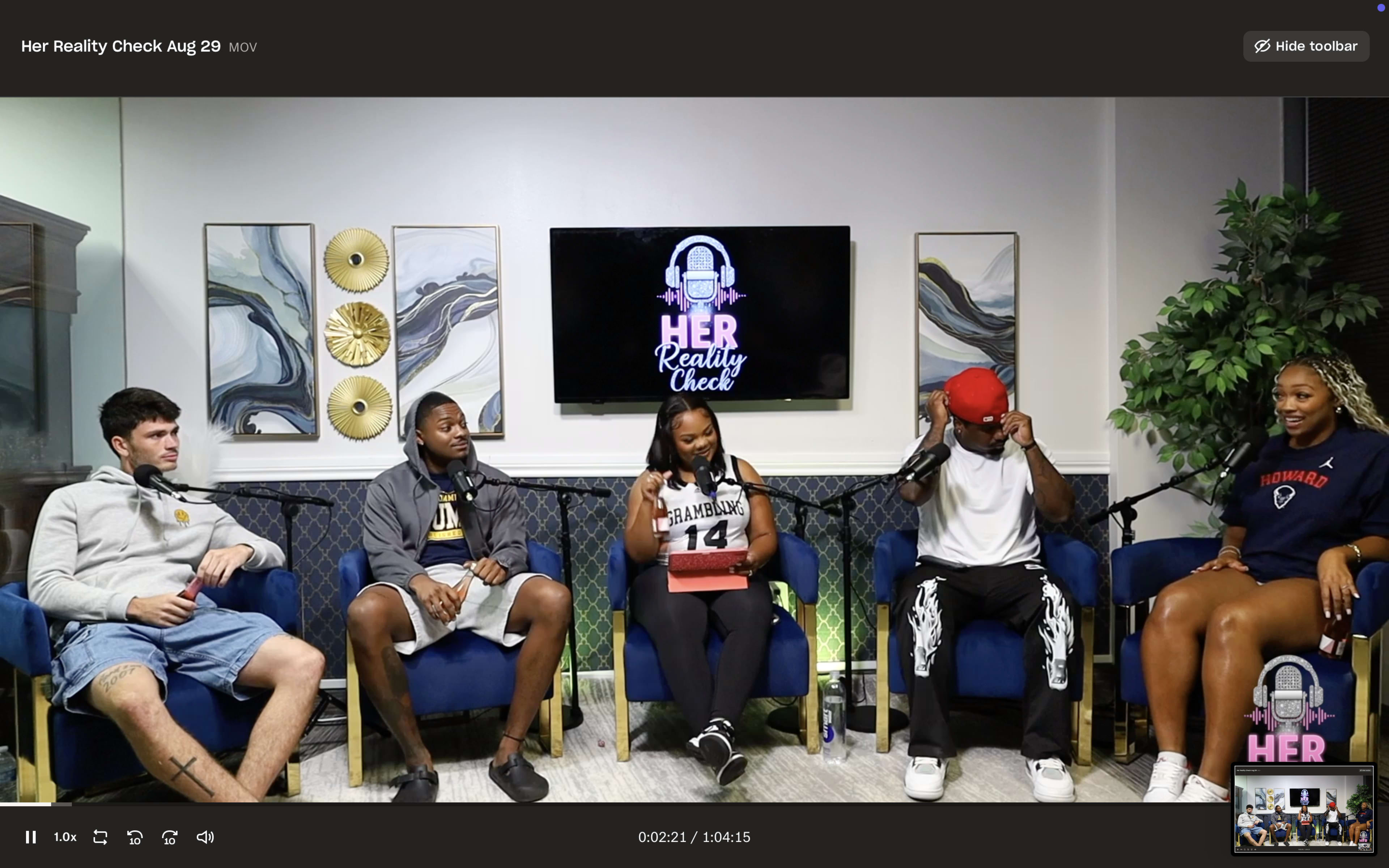 A group of five people sits in a studio setting with a large screen displaying the words "Her Reality Check" in the background.