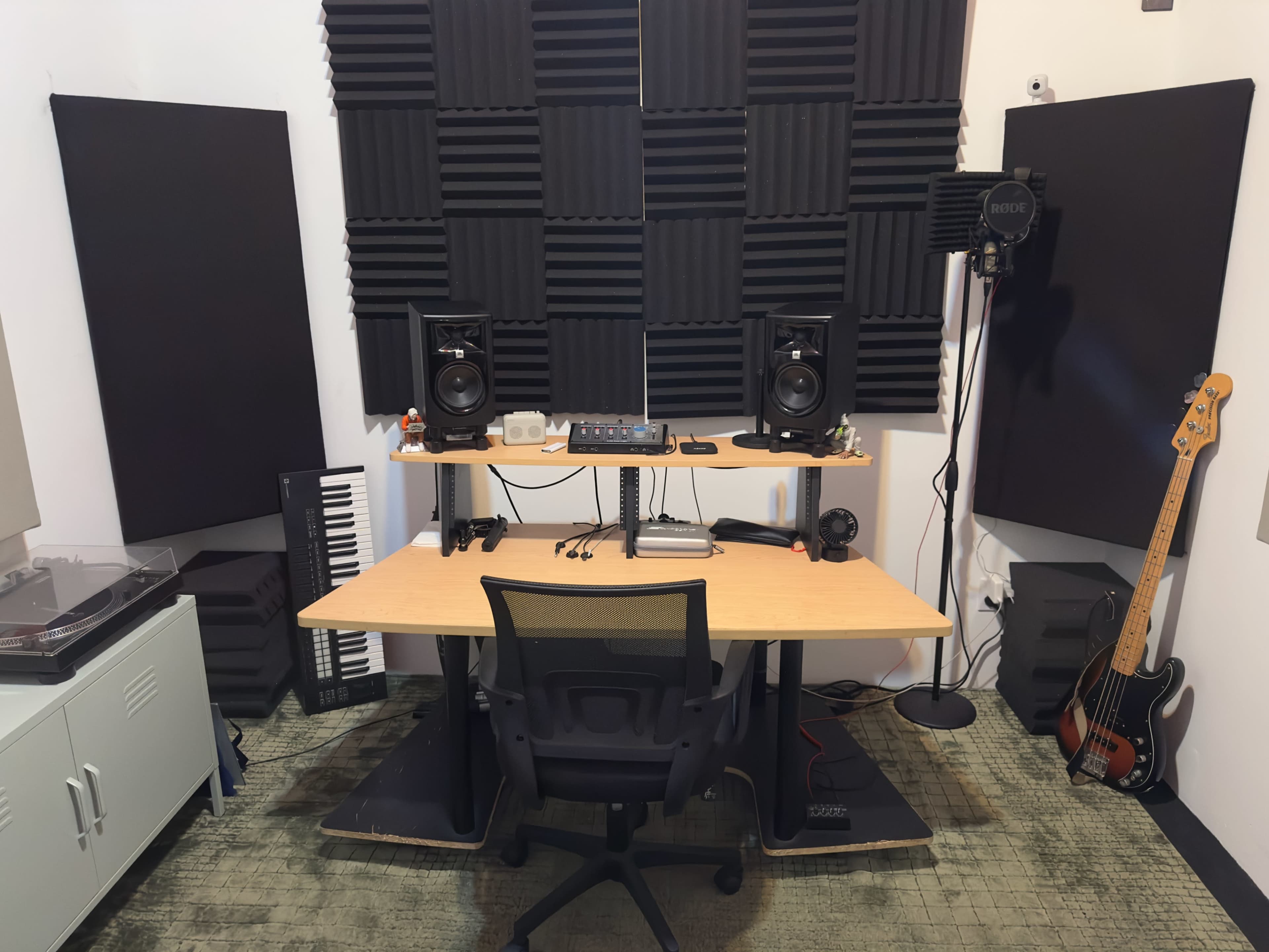 The image shows a music production studio setup featuring a wooden desk, studio monitors, a microphone, a keyboard, and various soundproofing panels on the walls.