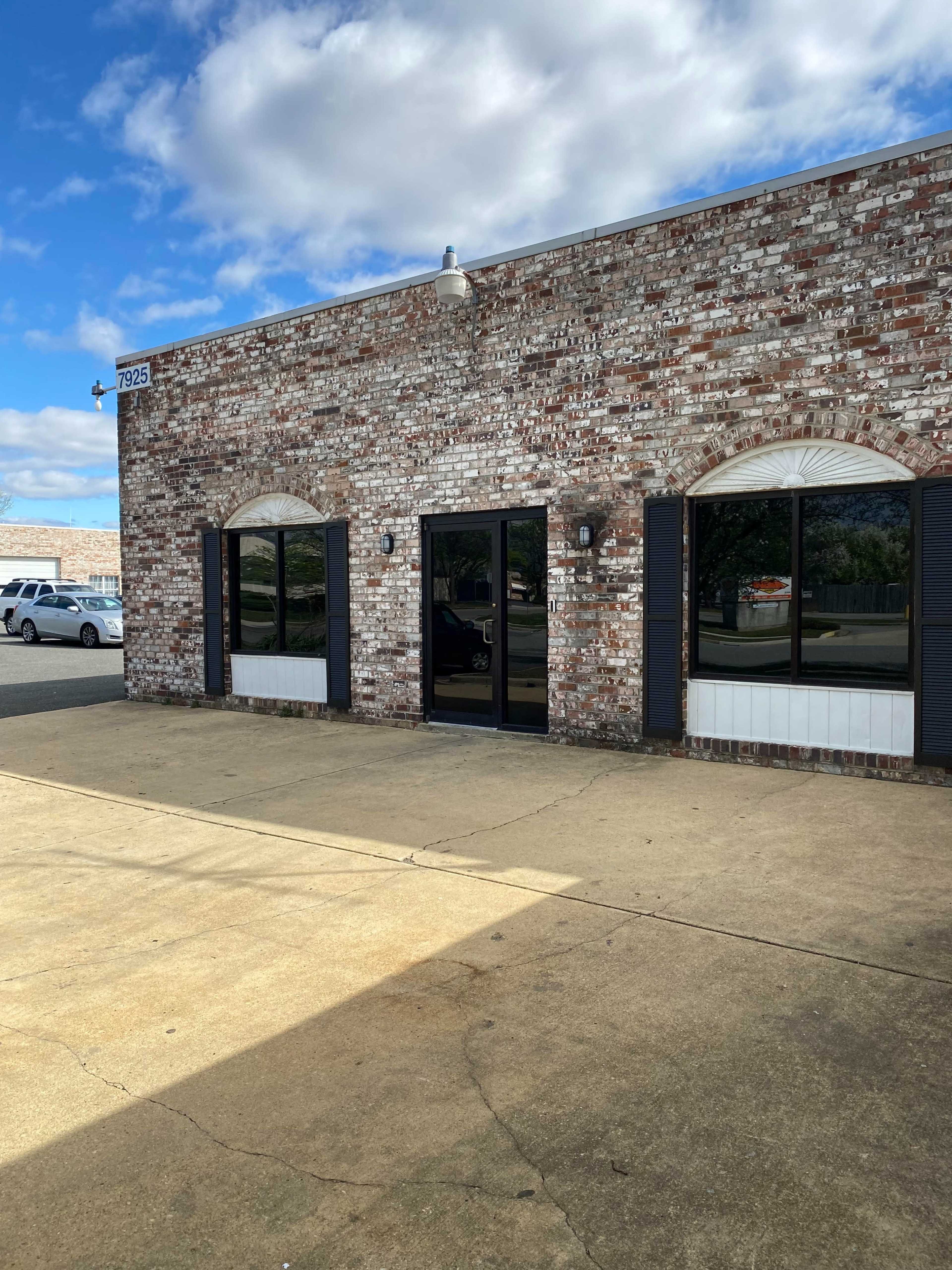 The image shows a brick building with large glass doors and window shutters, located on a paved area under a blue sky with scattered clouds.