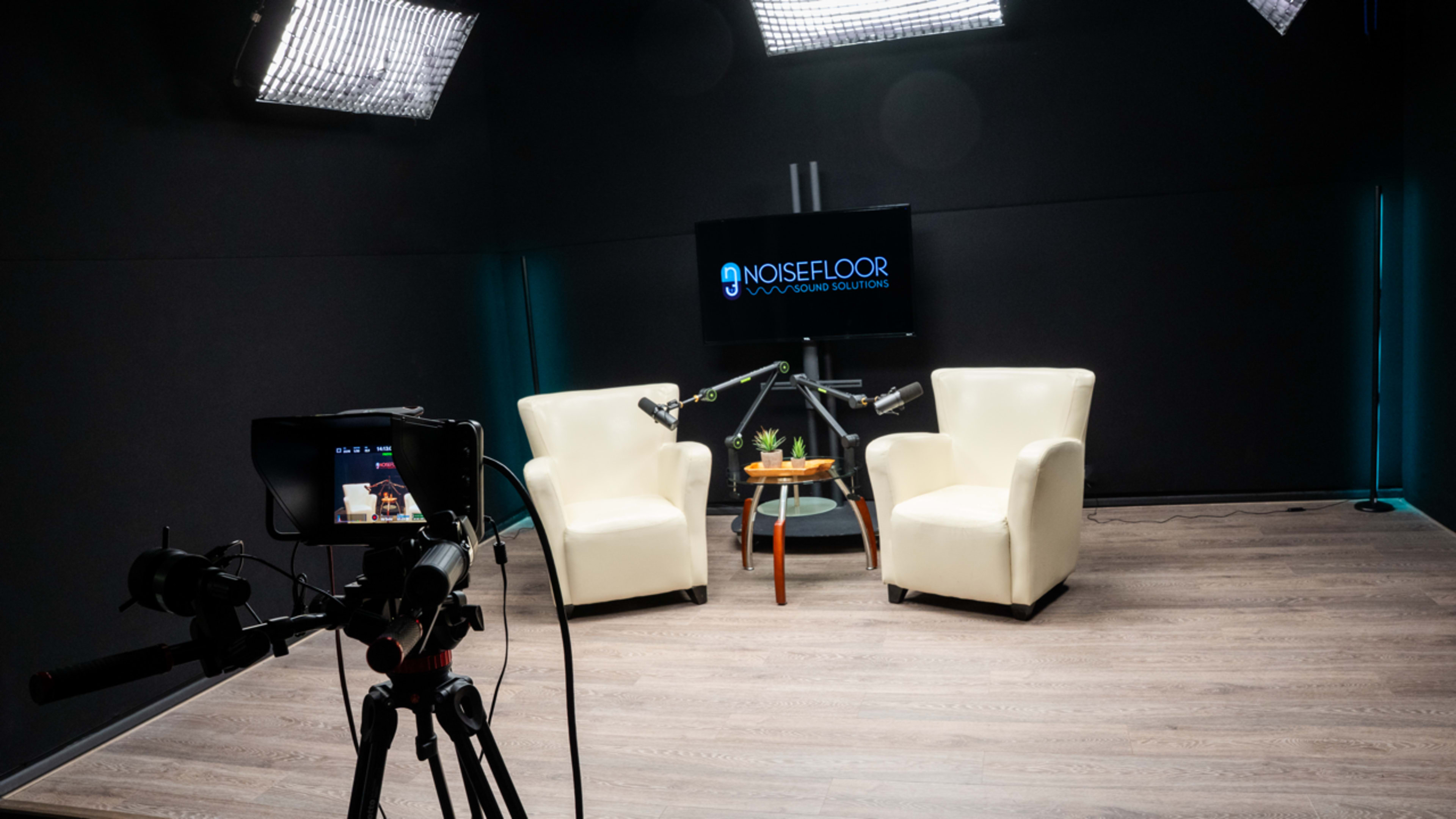 A well-lit studio setup features two white chairs, a small table with a plant, and cameras positioned to capture a television screen displaying the "NOISEFLOOR" logo.