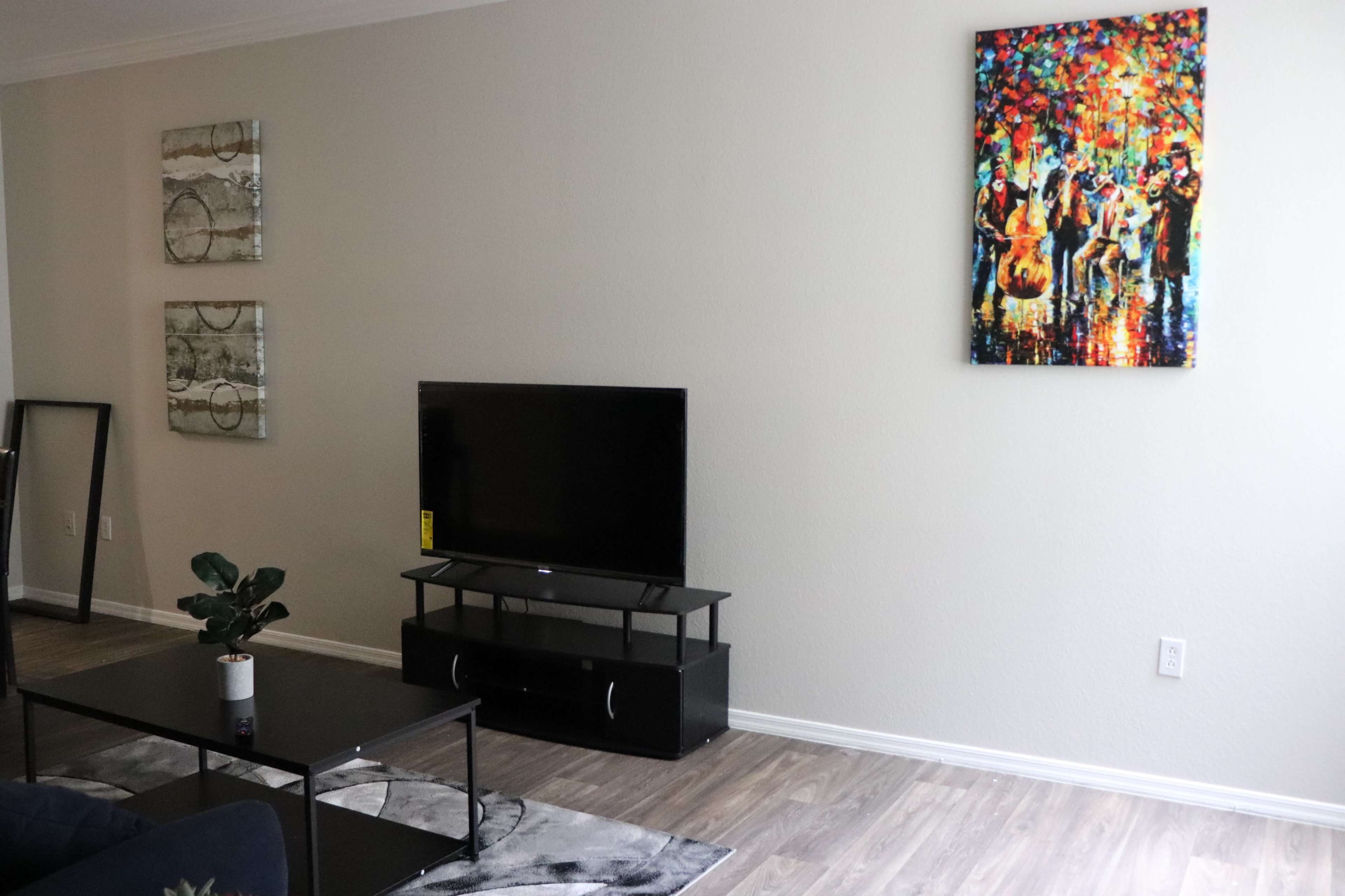 The image shows a living room with a television on a stand, a coffee table, and a colorful painting on the wall.