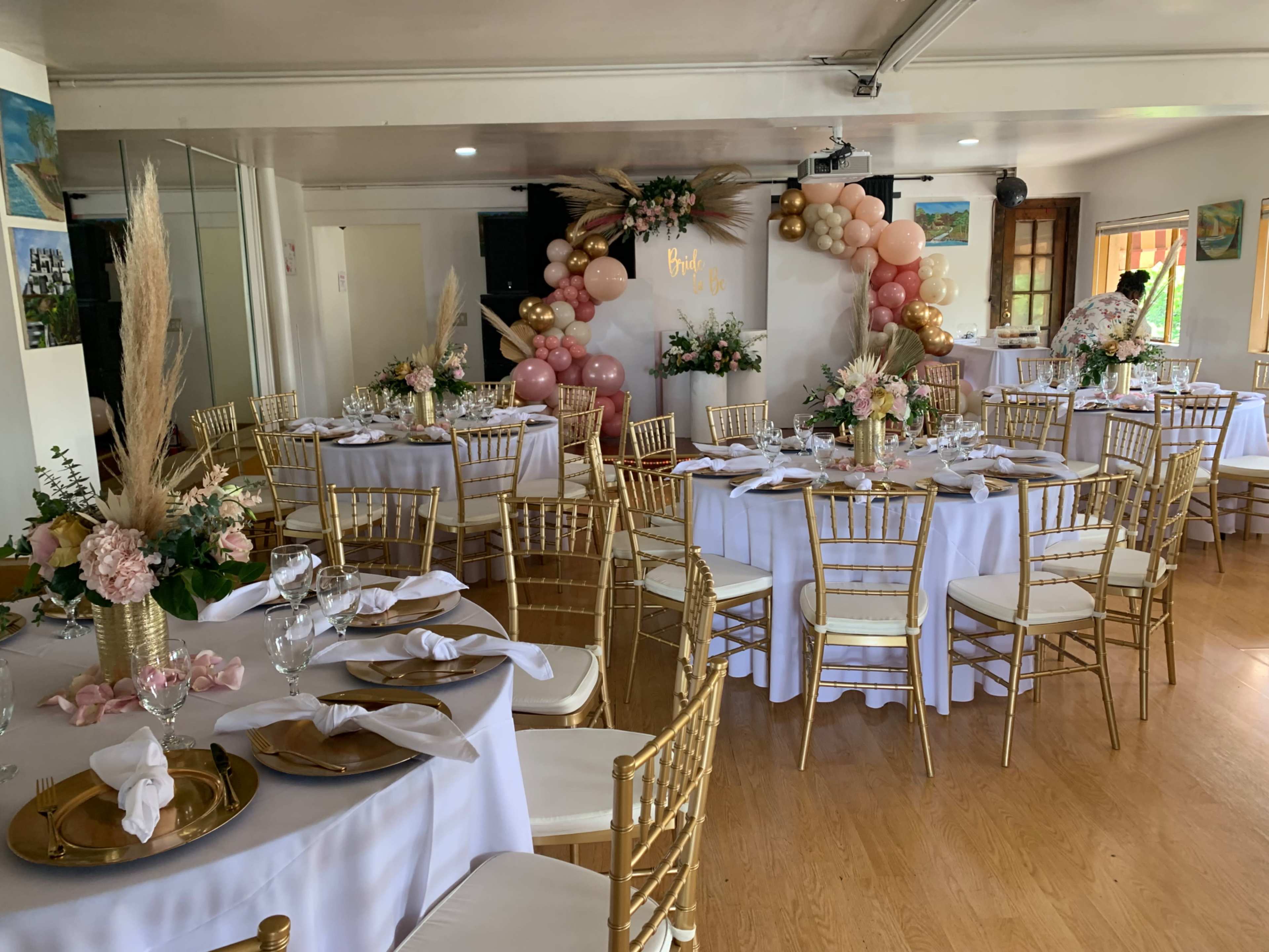 The image shows a decorated banquet hall with elegantly set round tables featuring white tablecloths, gold chairs, and centerpieces of flowers, alongside a backdrop adorned with balloons and greenery.