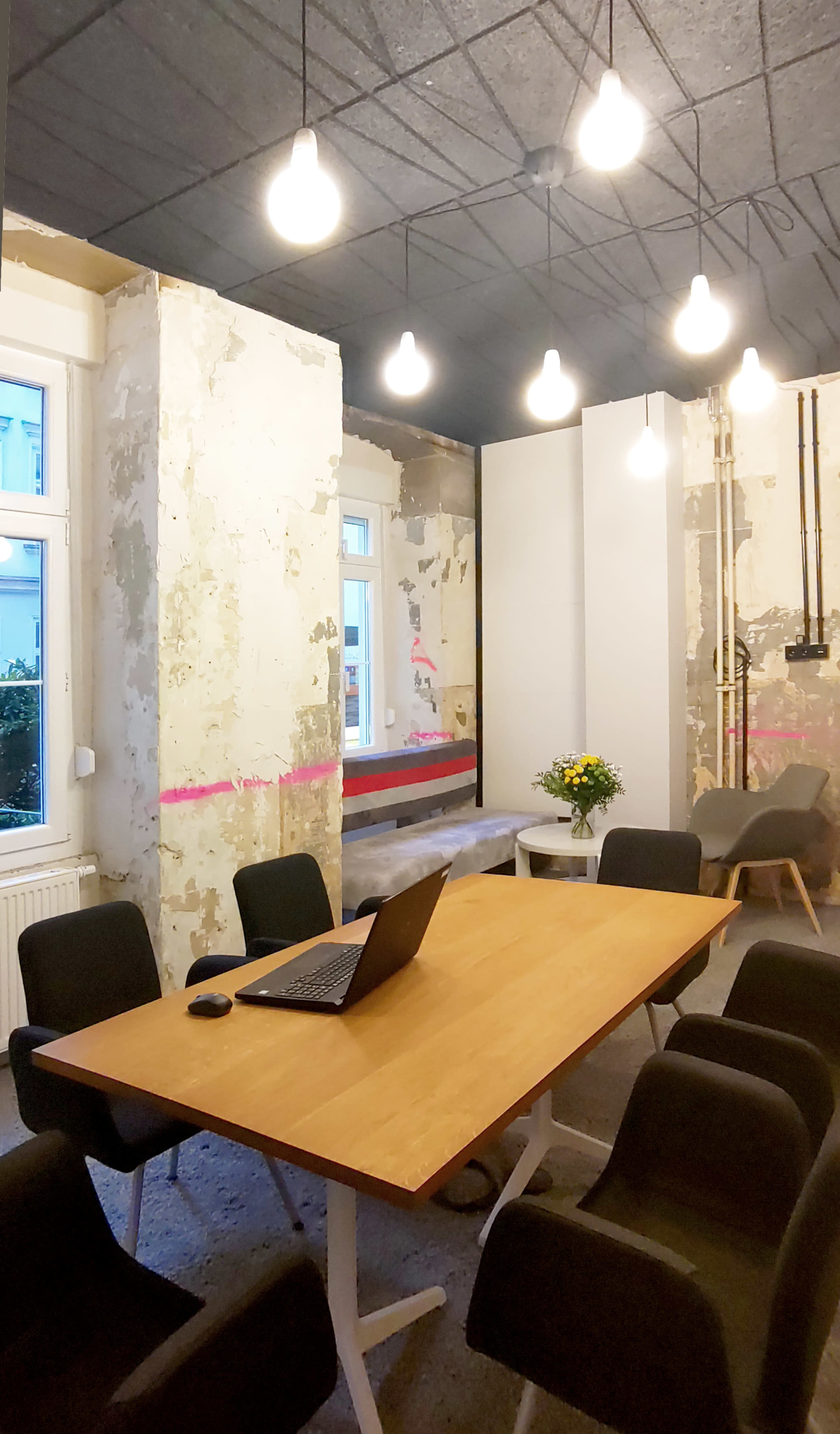 A meeting room features a wooden table with a laptop and a vase of flowers, surrounded by black chairs and unfinished walls with exposed drywall.