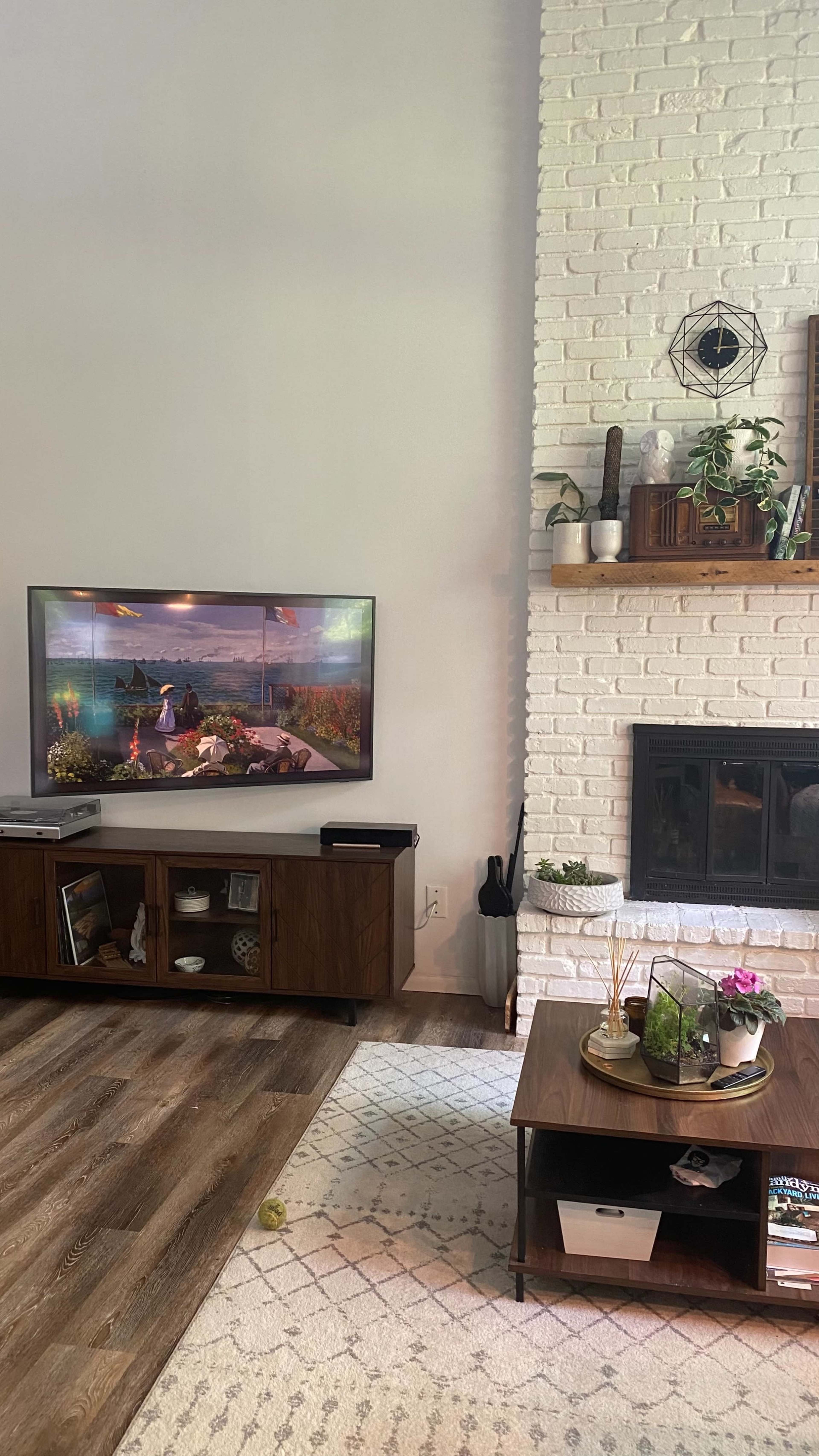 A living room with a television mounted on the wall, a wooden entertainment unit beneath it, and a white brick fireplace with decorative items and plants on the mantel.
