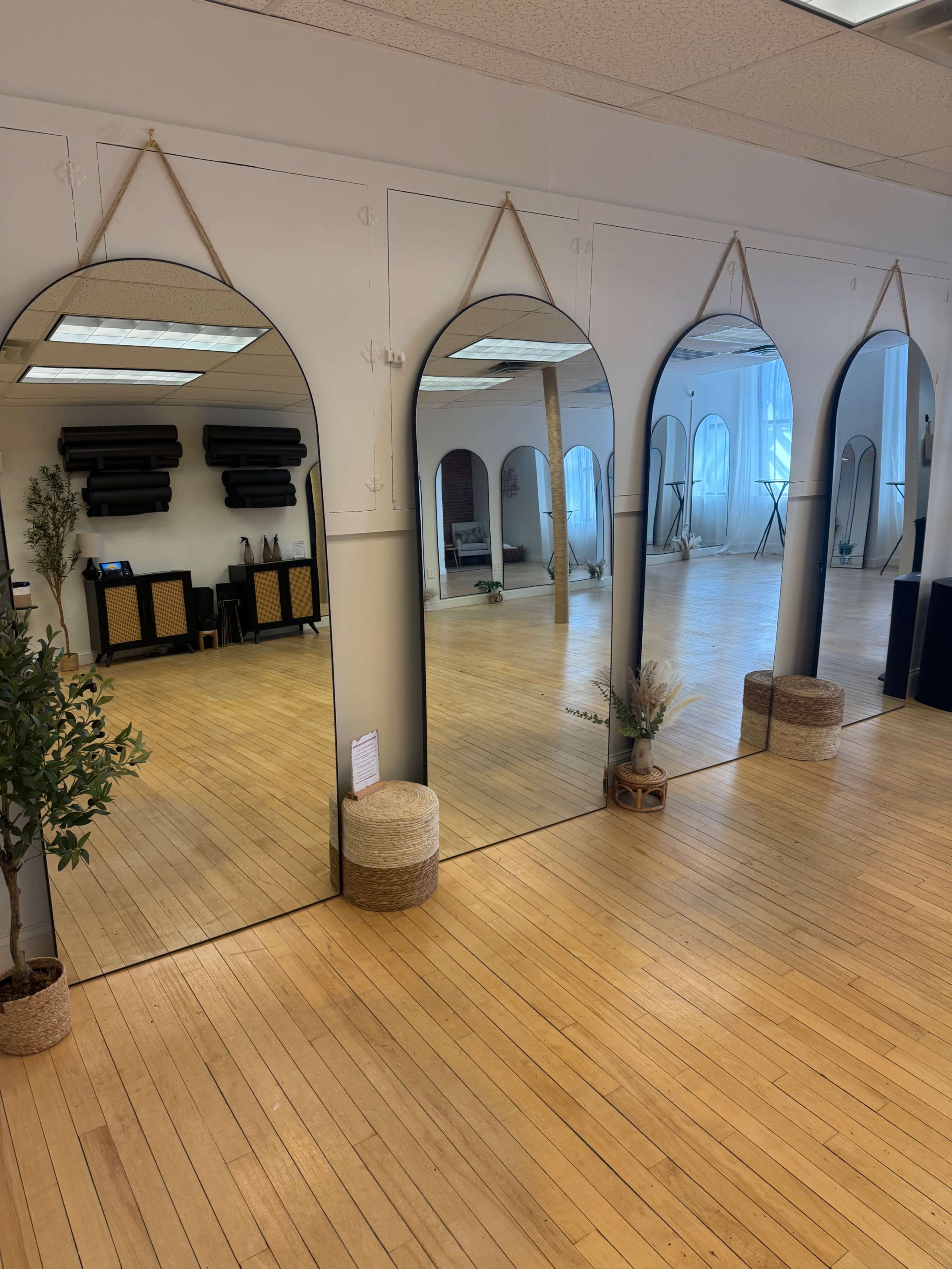The image shows a spacious room with wooden floors featuring three tall, arched mirrors lined up against a wall, accompanied by decorative plants and woven baskets.