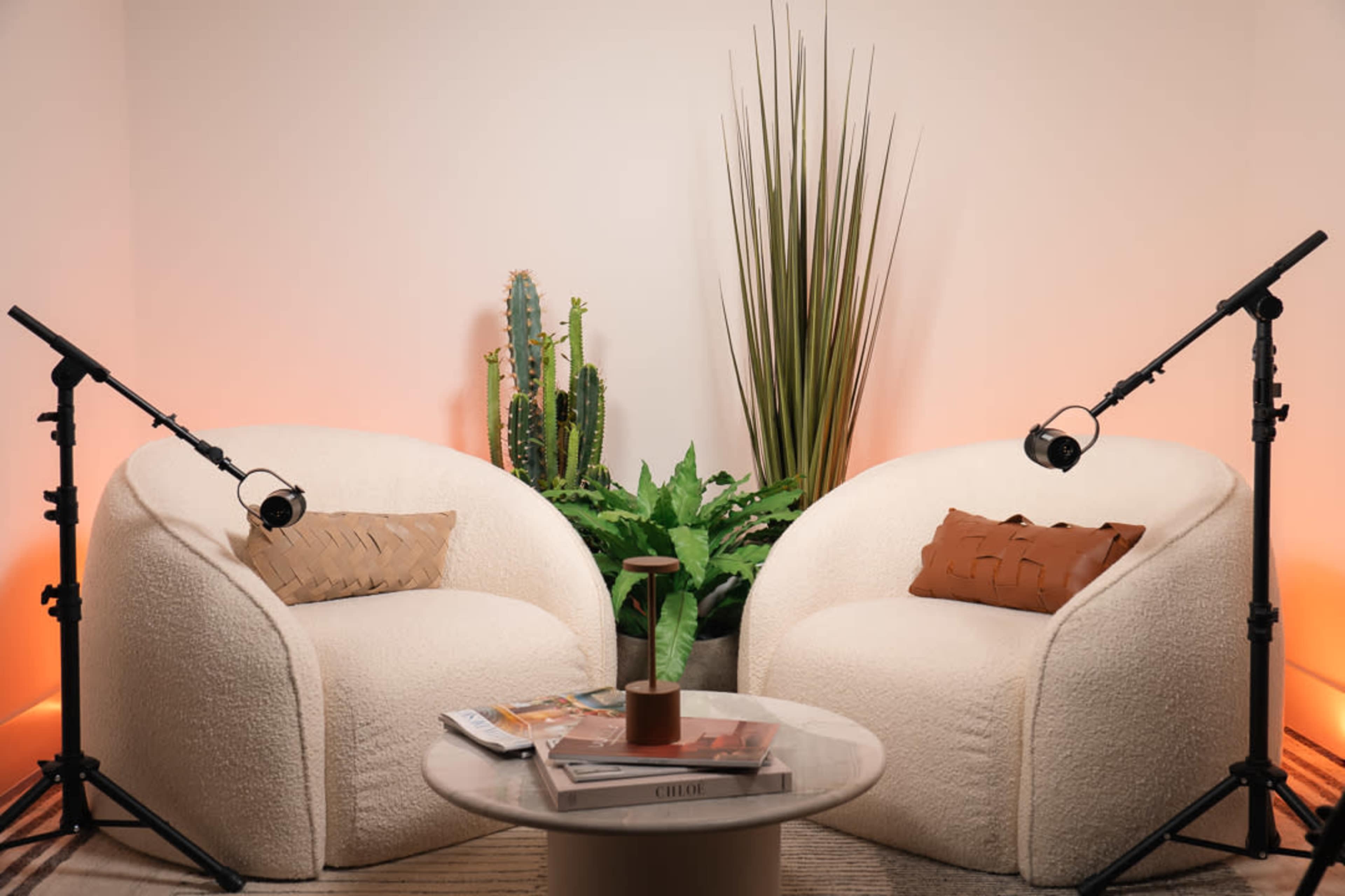 The image features two plush white chairs facing each other, accompanied by a round table decorated with magazines, and surrounded by greenery and soft lighting.