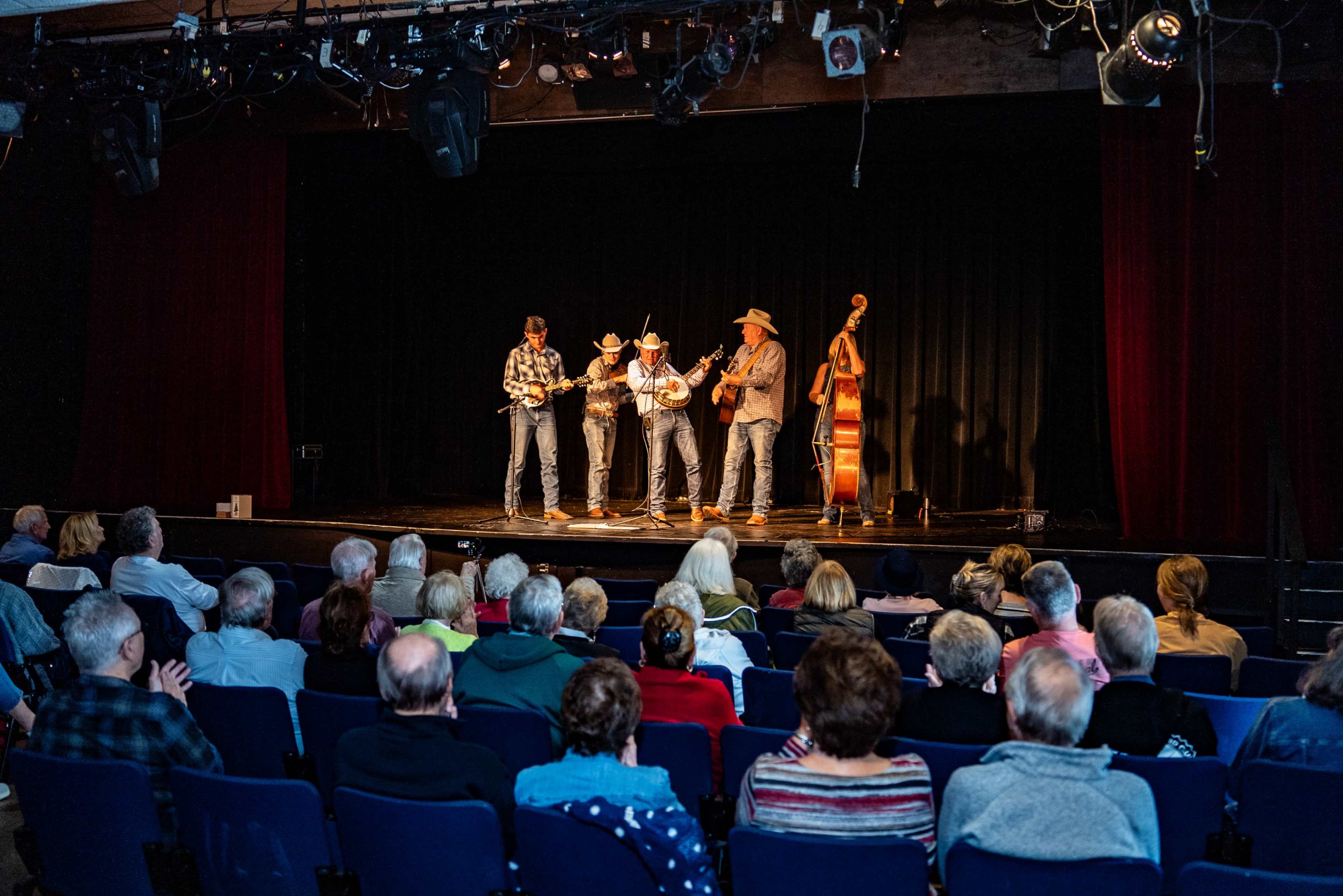 A bluegrass band performs on stage for an audience seated in a theater.