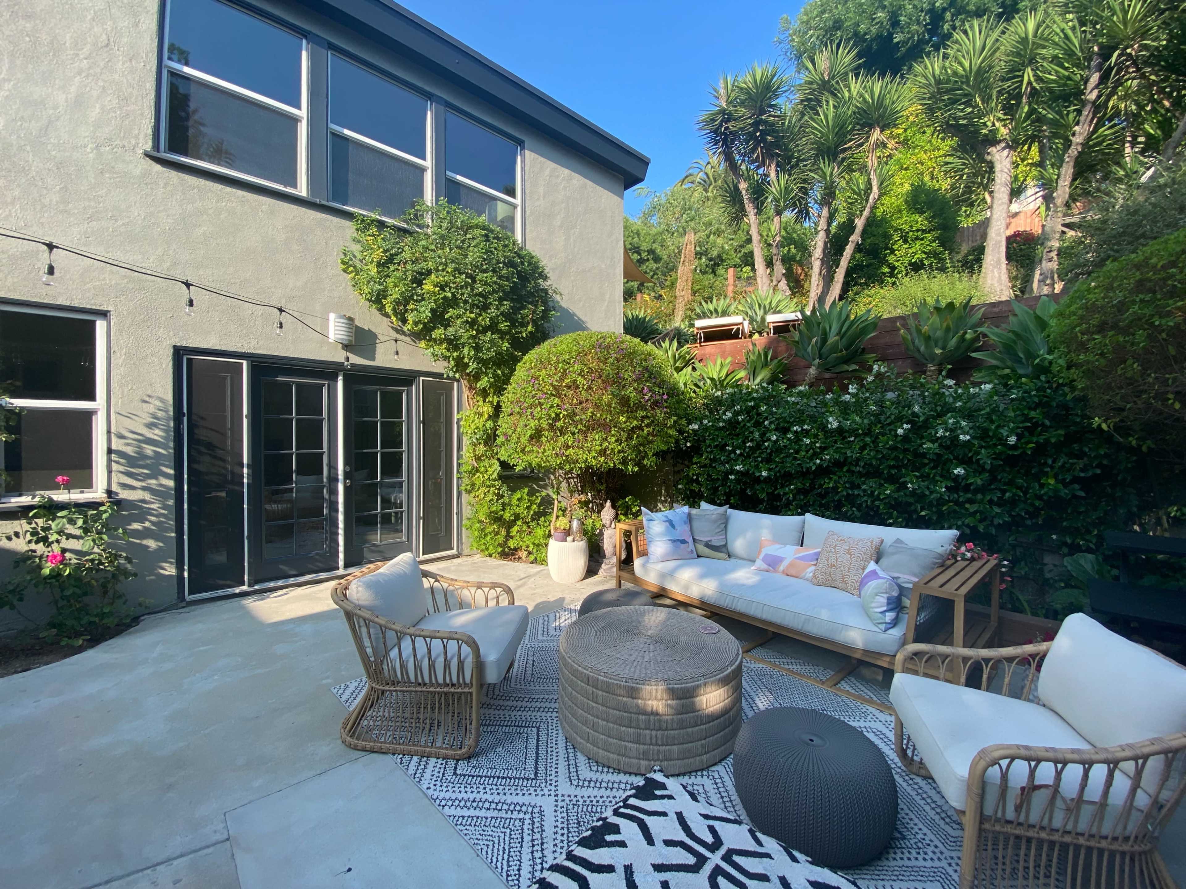 The image shows a cozy outdoor seating area with a white sofa, two wicker chairs, and a round coffee table surrounded by greenery and a gray building.