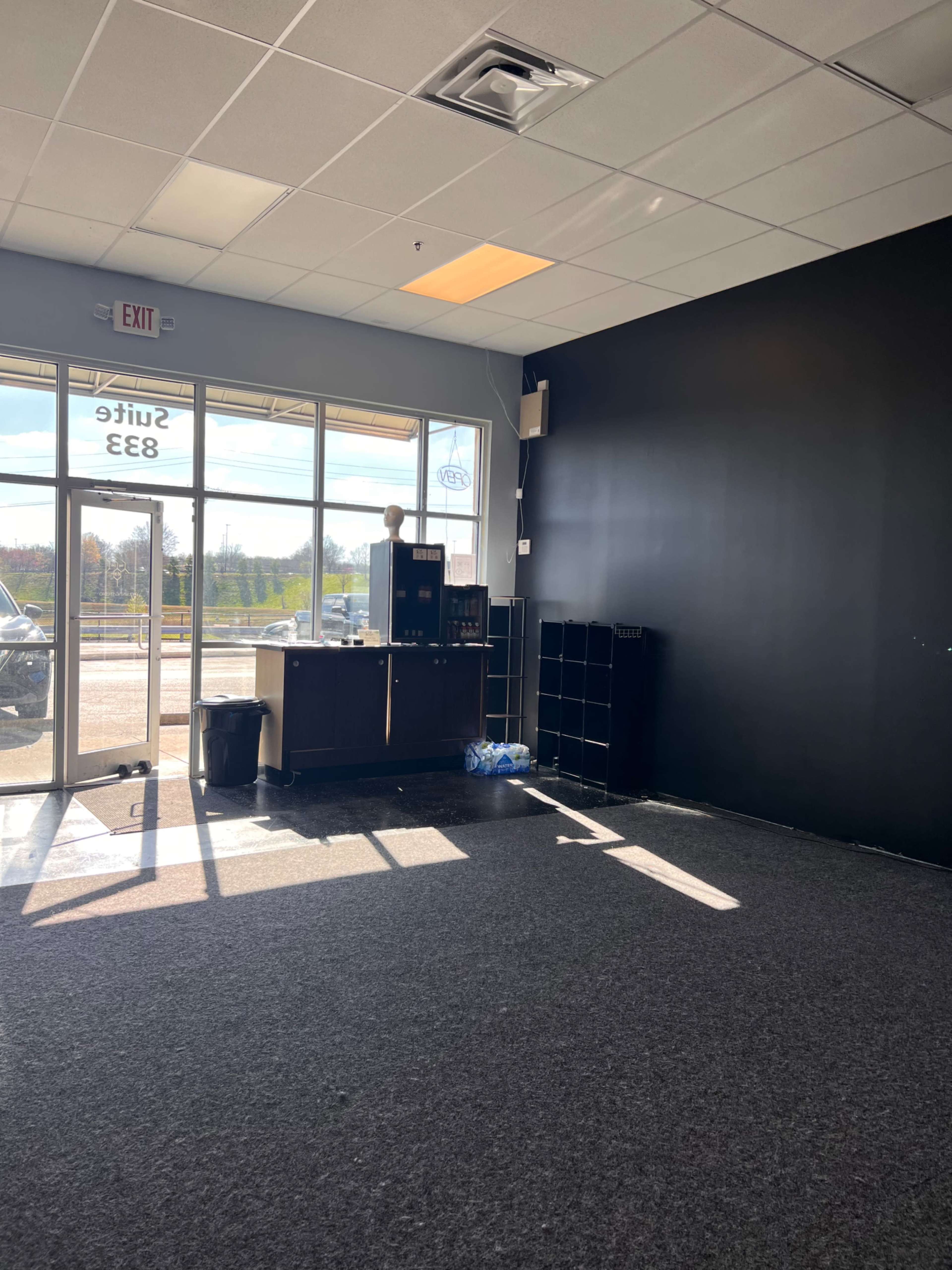 A vacant commercial space with a dark wall, a large window allowing natural light, and minimal furnishings including a counter and black shelving units.
