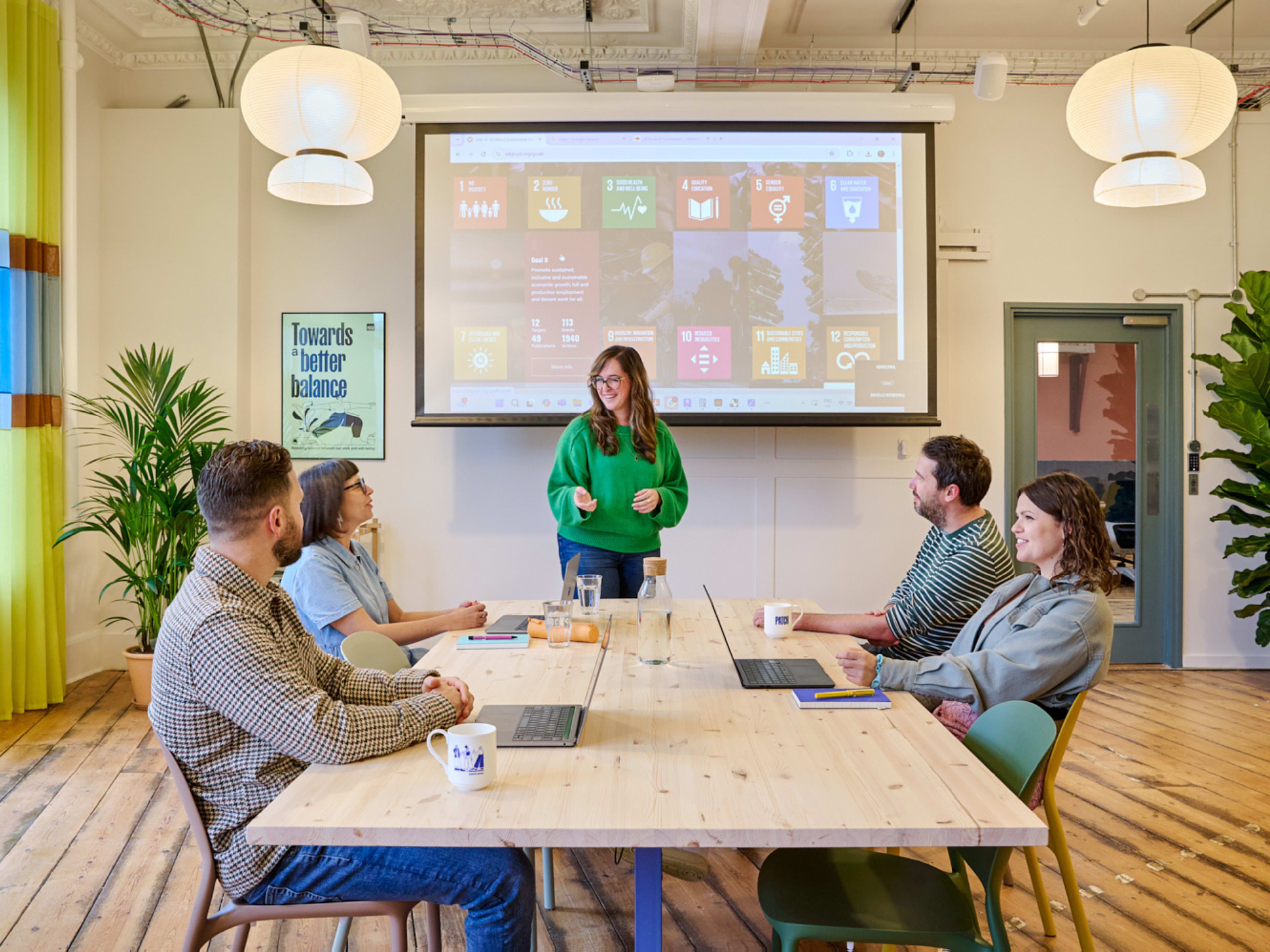 A woman stands at the front of a modern meeting room presenting to four colleagues seated at a long table, with a projector screen displaying various digital content in the background.
