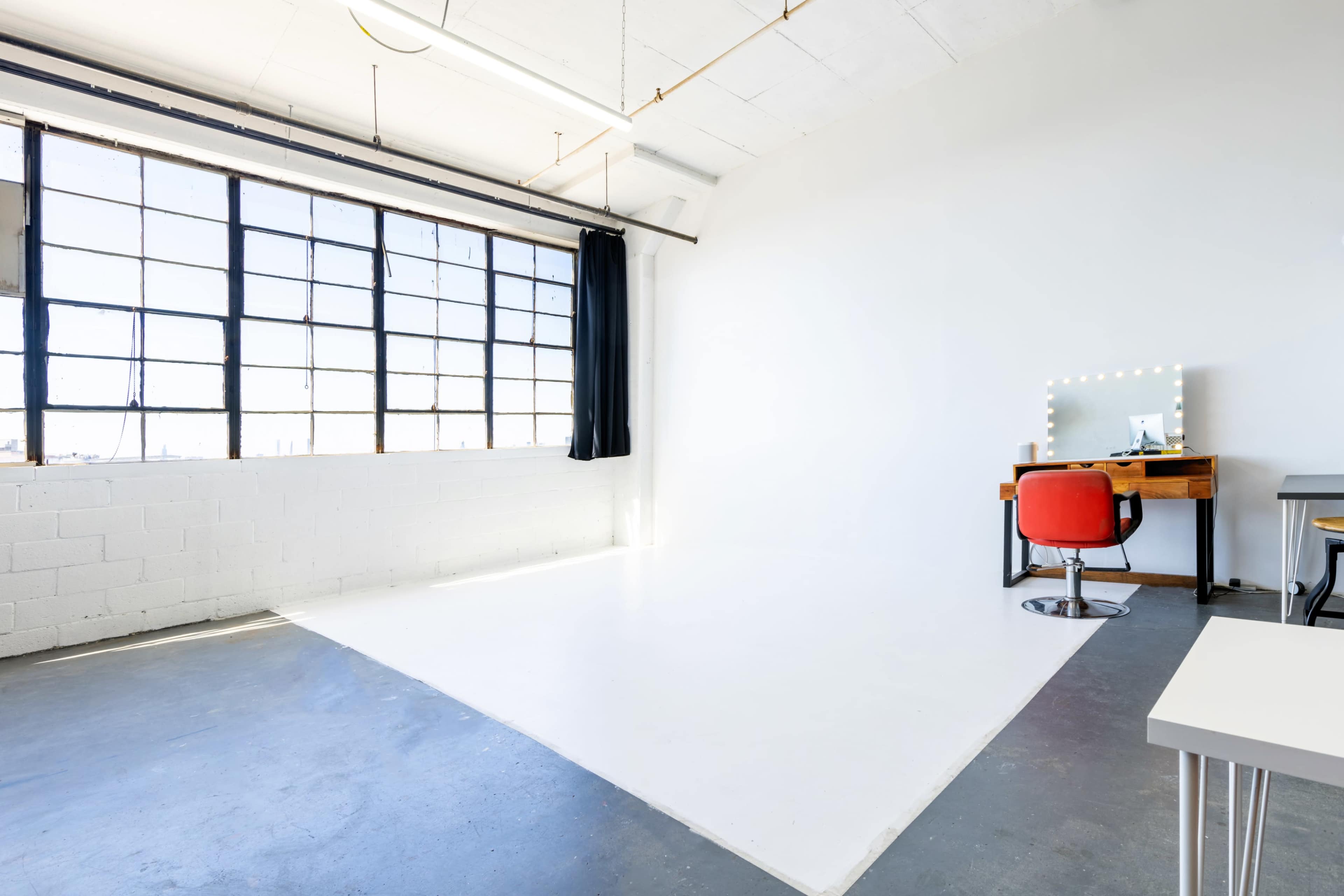 A bright, empty studio features a large window, a red chair at a wooden desk with a mirror, and a simple white wall.