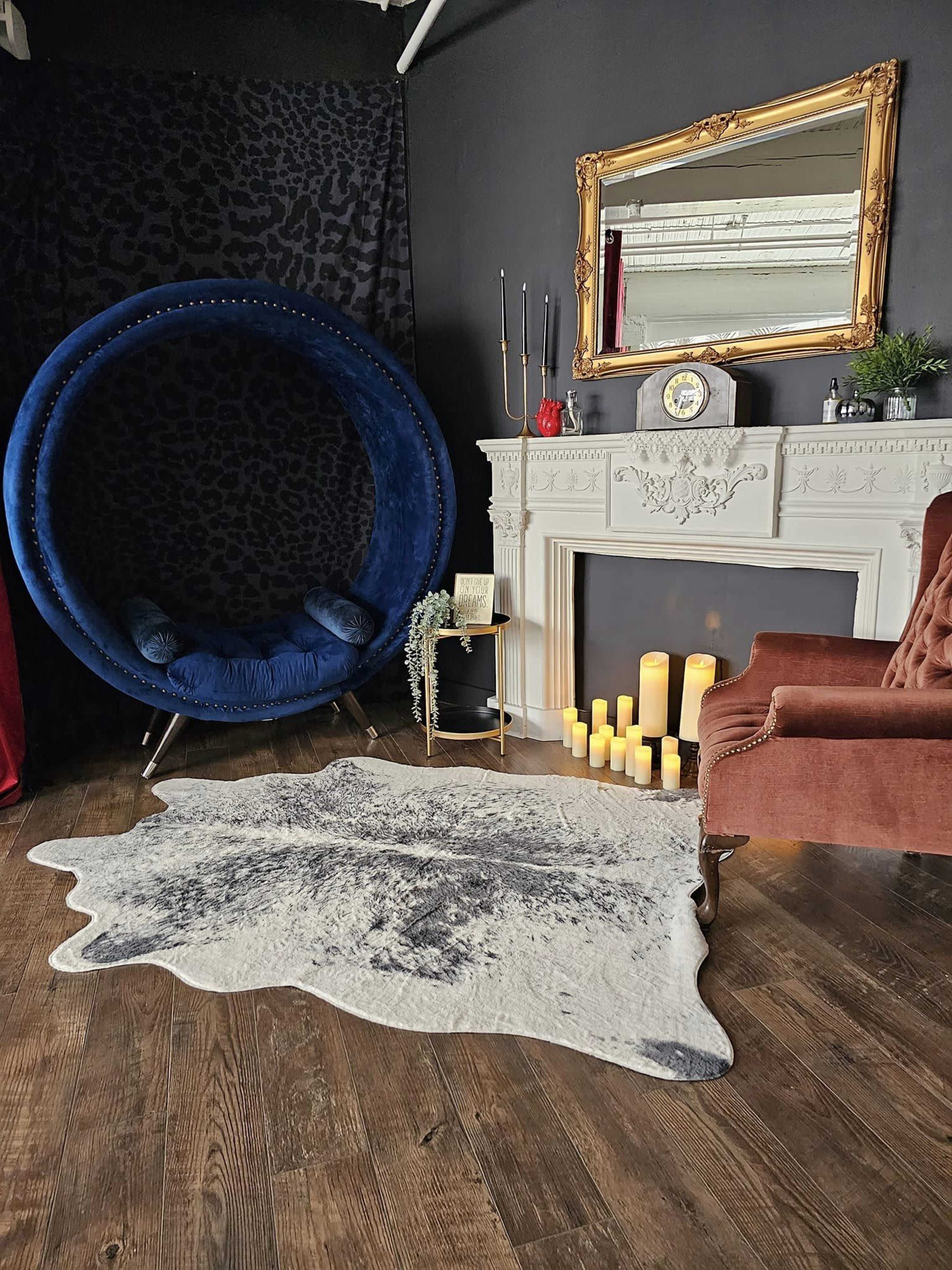 The image shows a stylish interior with a large blue circular chair, a fireplace adorned with candles, a gold-framed mirror, and a cowhide rug on a wooden floor.