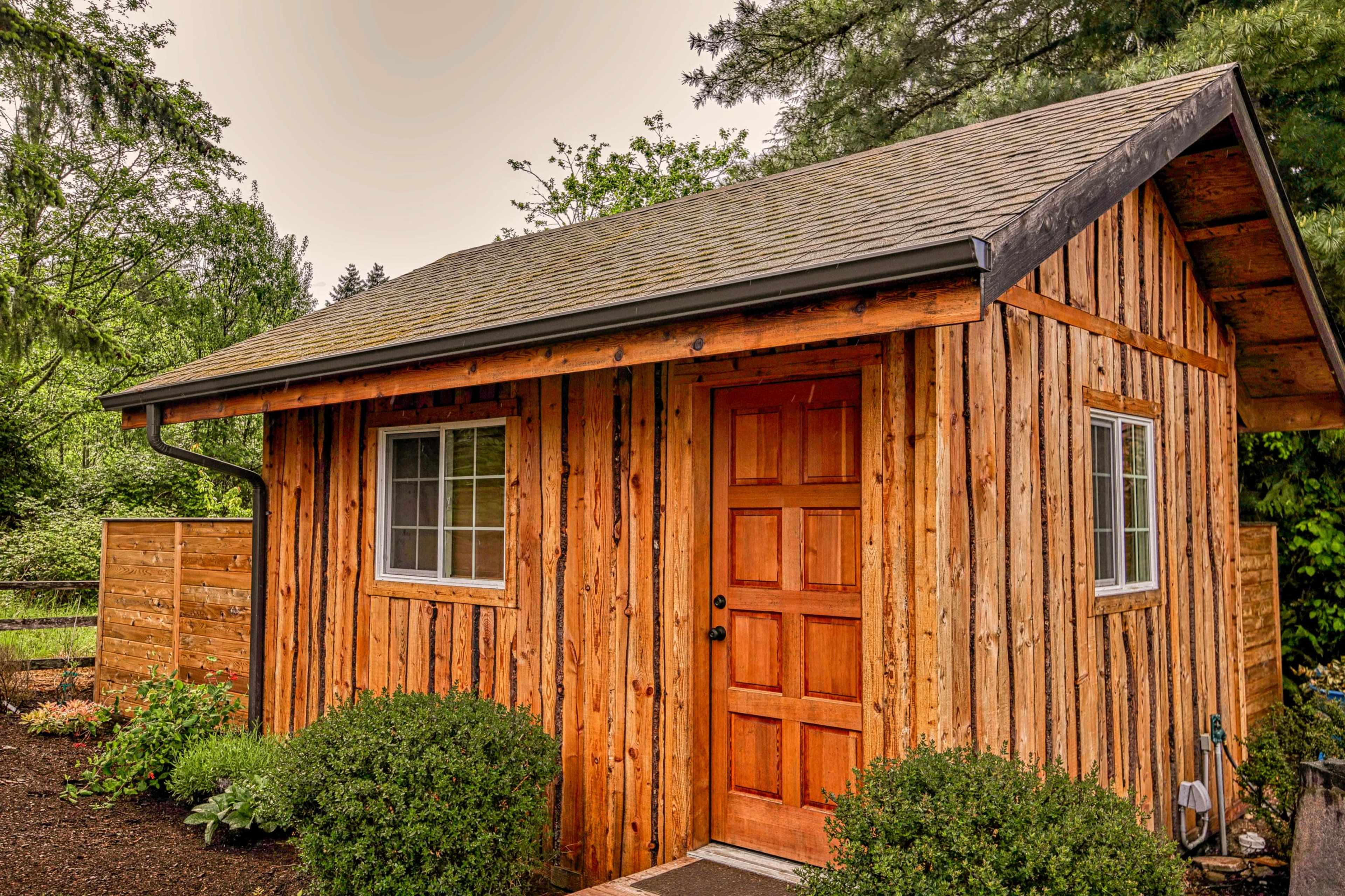 A small wooden cabin with a brown door and two windows, surrounded by neatly trimmed bushes and trees.