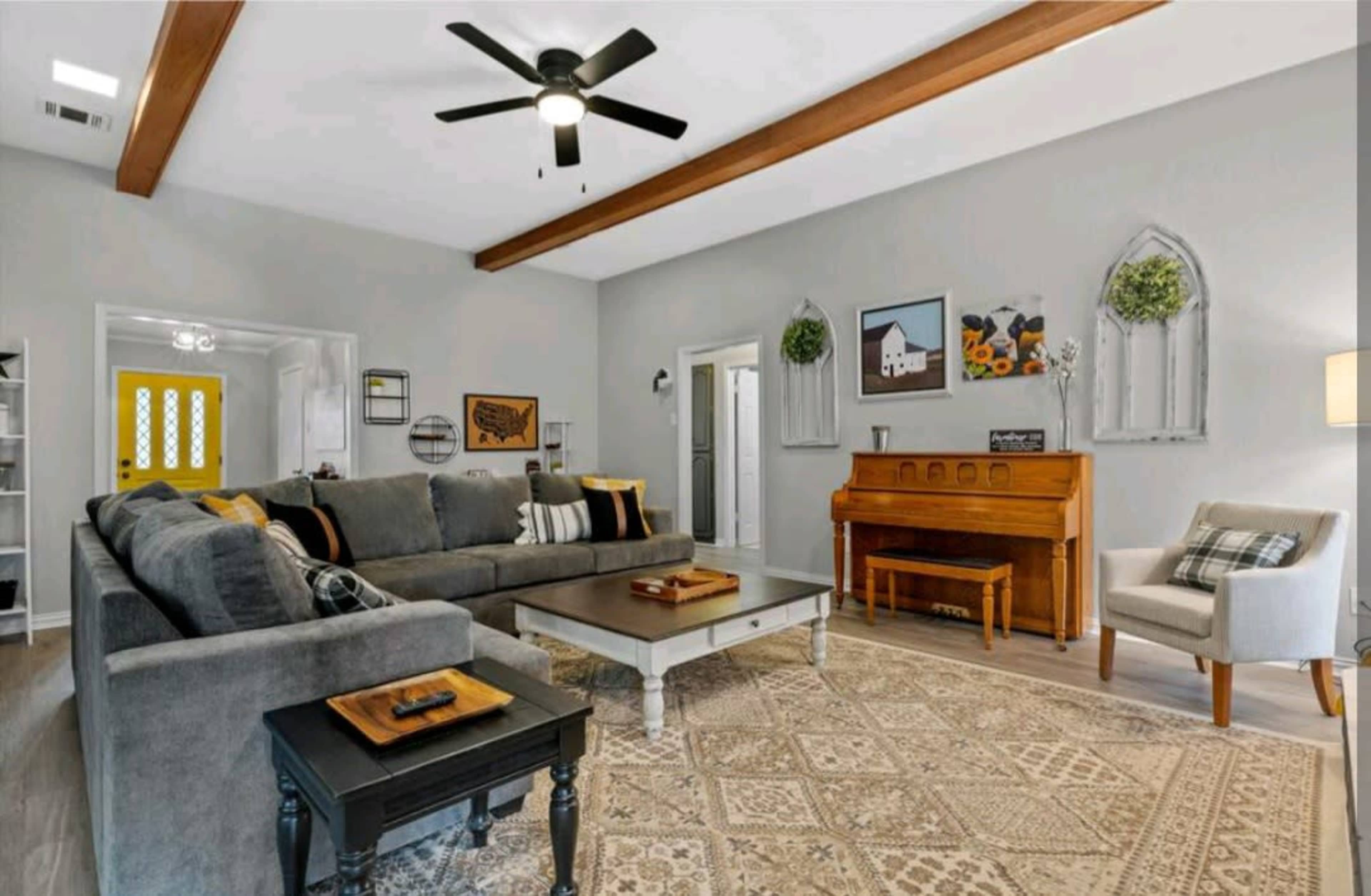 The living room features a large sectional sofa, a wooden piano, a coffee table, and a decorative area rug beneath a ceiling fan with exposed beams.