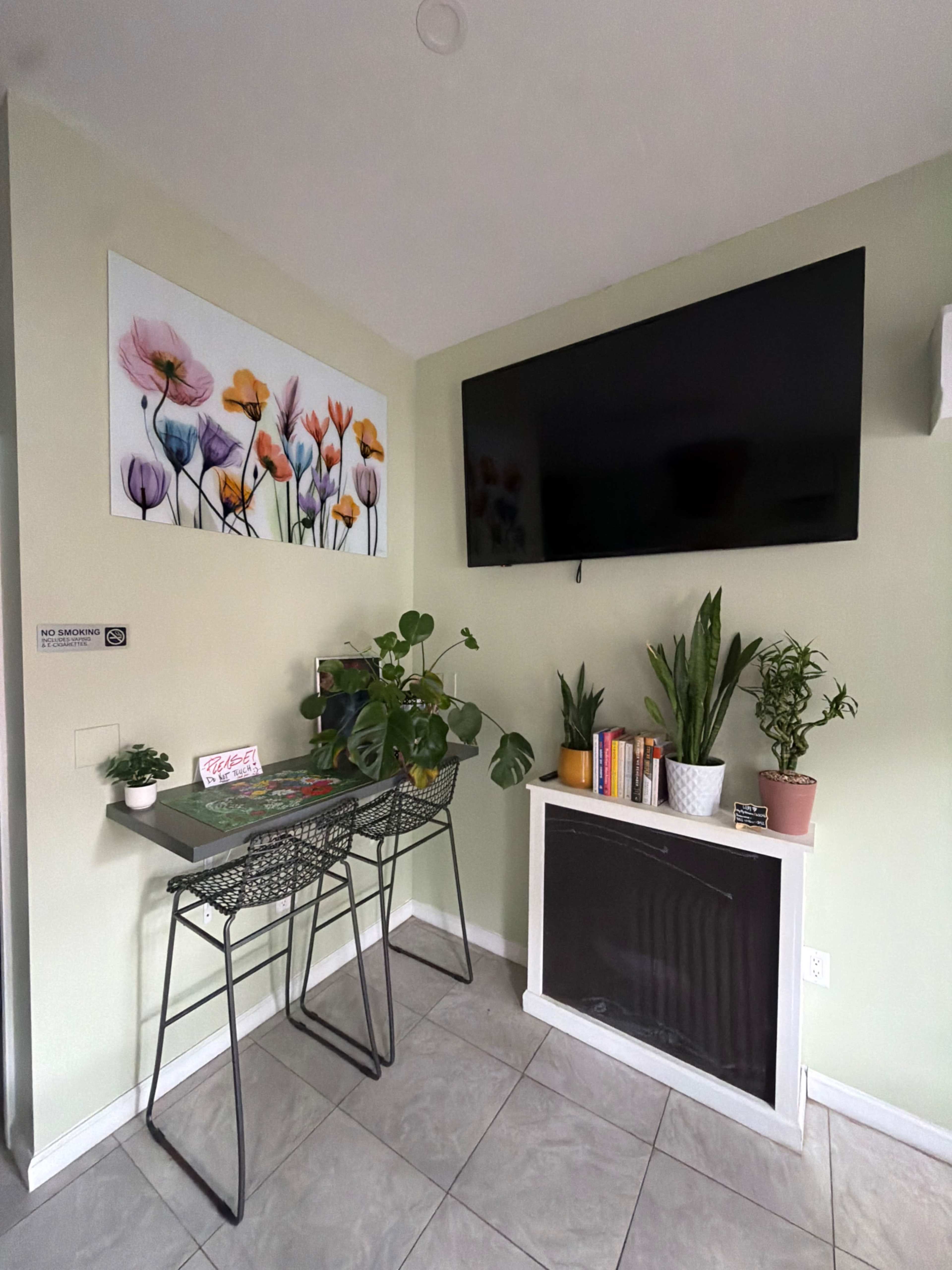 The image shows a small kitchen area with a wall-mounted TV, a decorative floral artwork, a table with two stools, and several potted plants.
