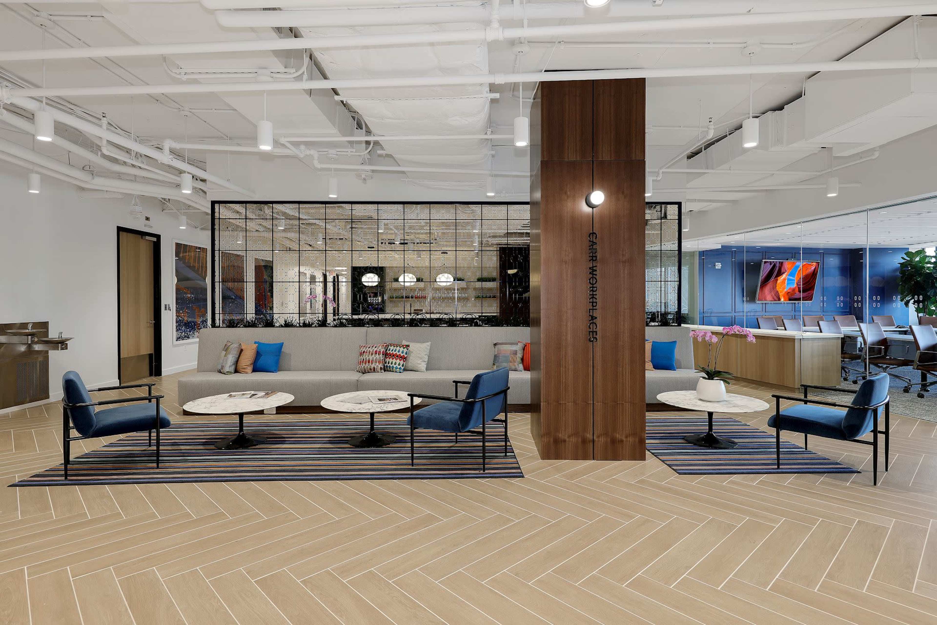 The image shows a modern office lobby with a seating area featuring a gray sofa and blue chairs, surrounded by a wooden column and large glass windows.