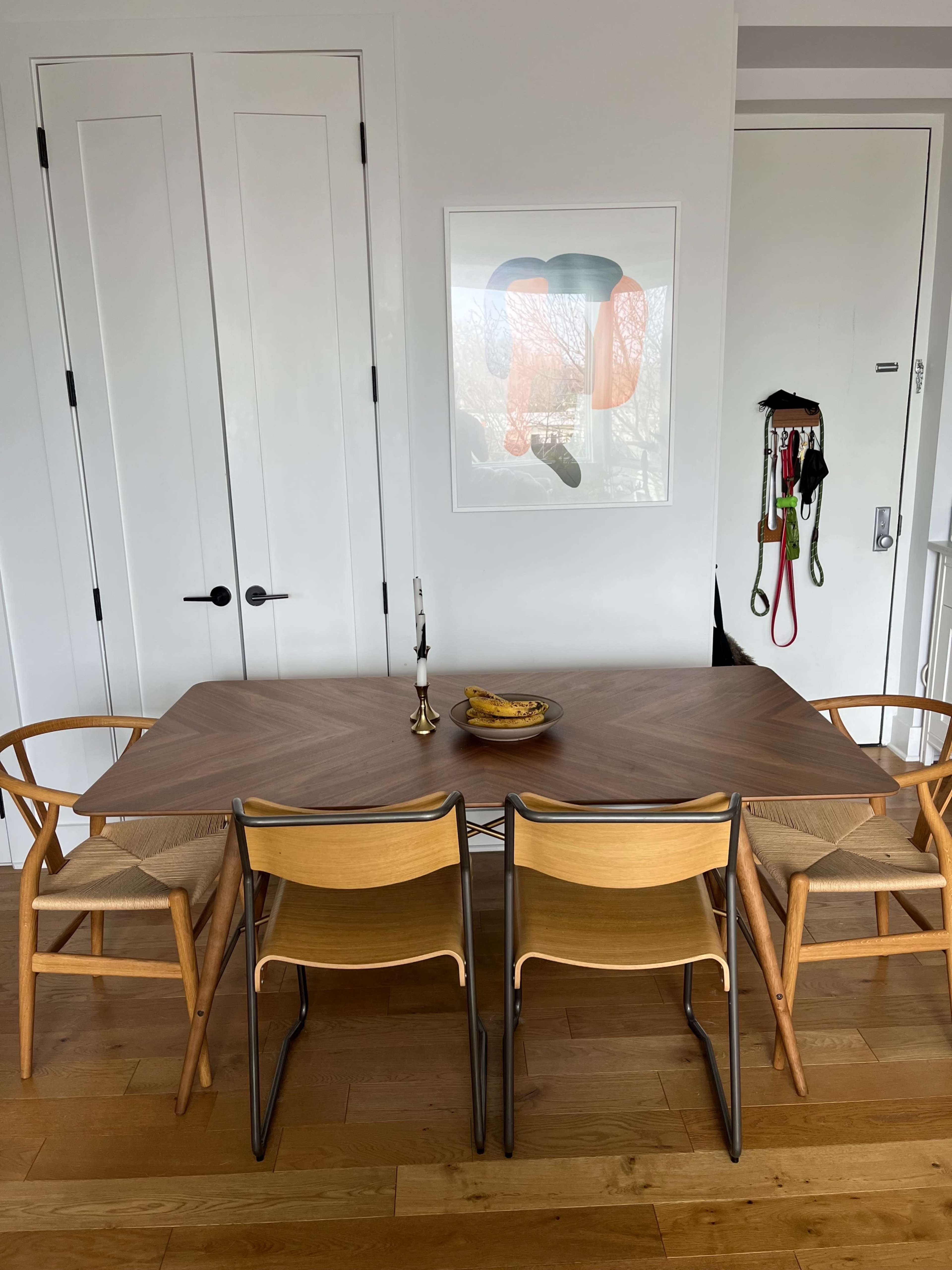 A dining table with wooden chairs and a candle holder is set against a wall featuring a framed artwork and an open door.
