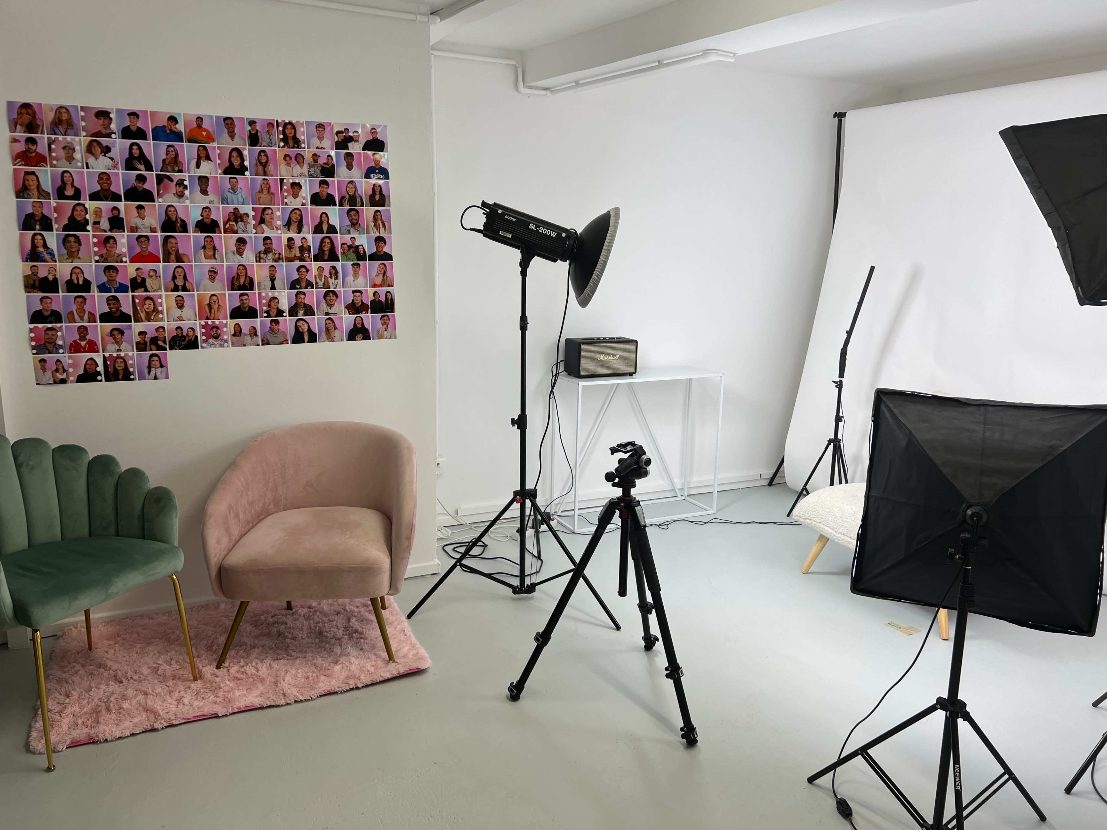 The image shows a photography studio setup with a wall displaying a collage of portraits, two chairs, lighting equipment, and a camera on a tripod.
