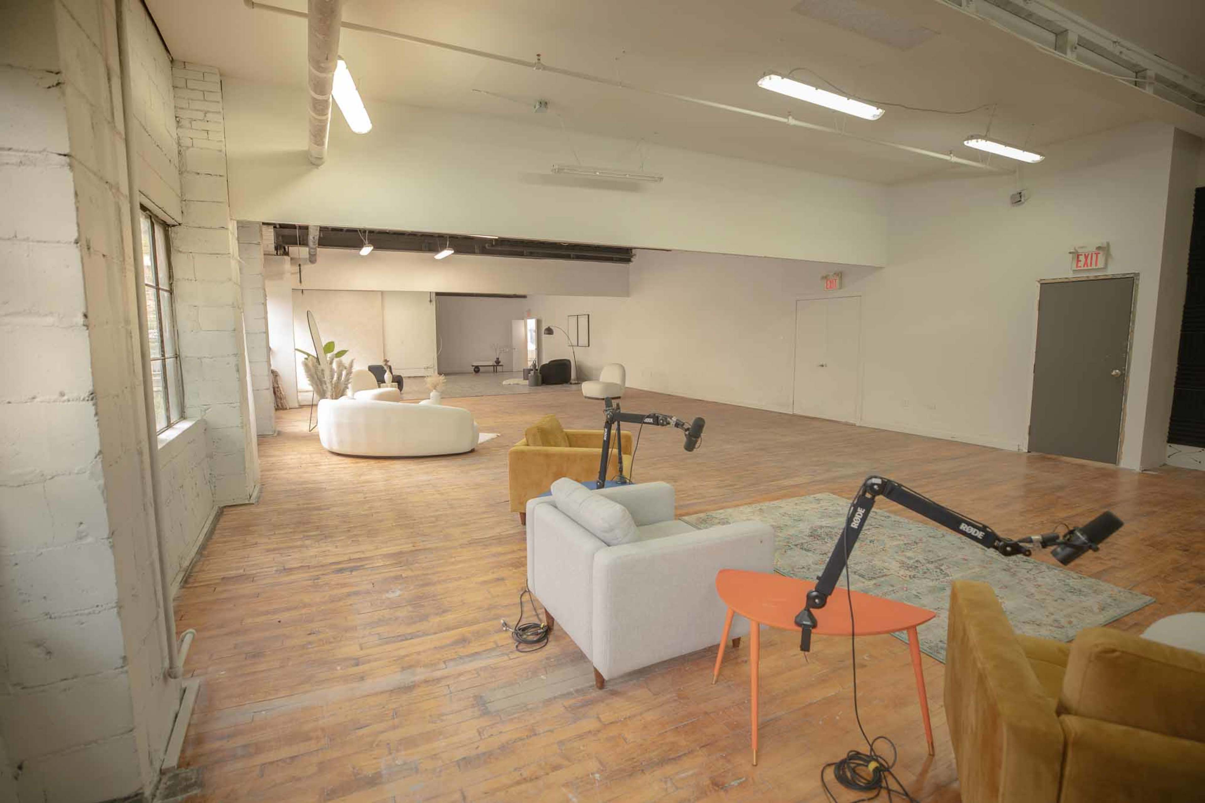 The image shows a spacious, modern interior with wooden flooring, a white sofa, yellow armchairs, and adjustable lamps, along with large windows allowing natural light.