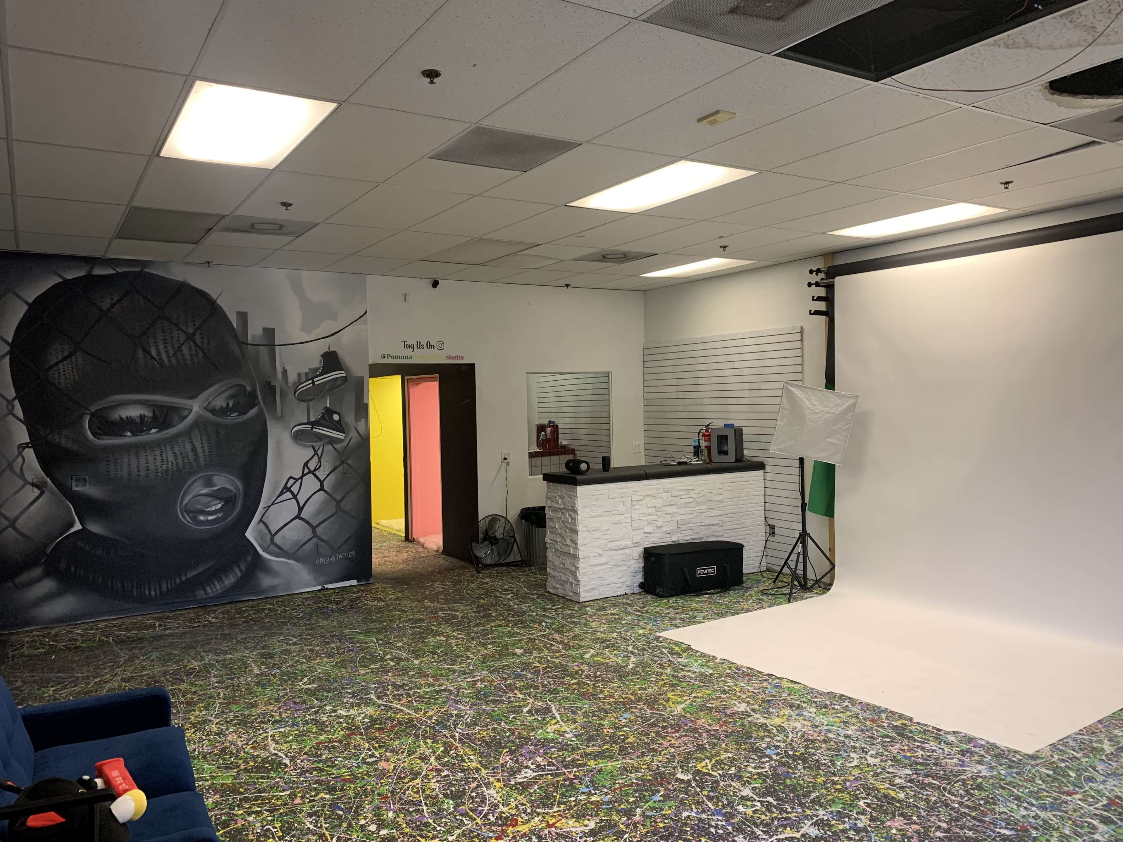 The image shows an indoor studio space with a graffiti mural of a masked figure on one wall, a white backdrop on a stand, and a colorful painted floor.