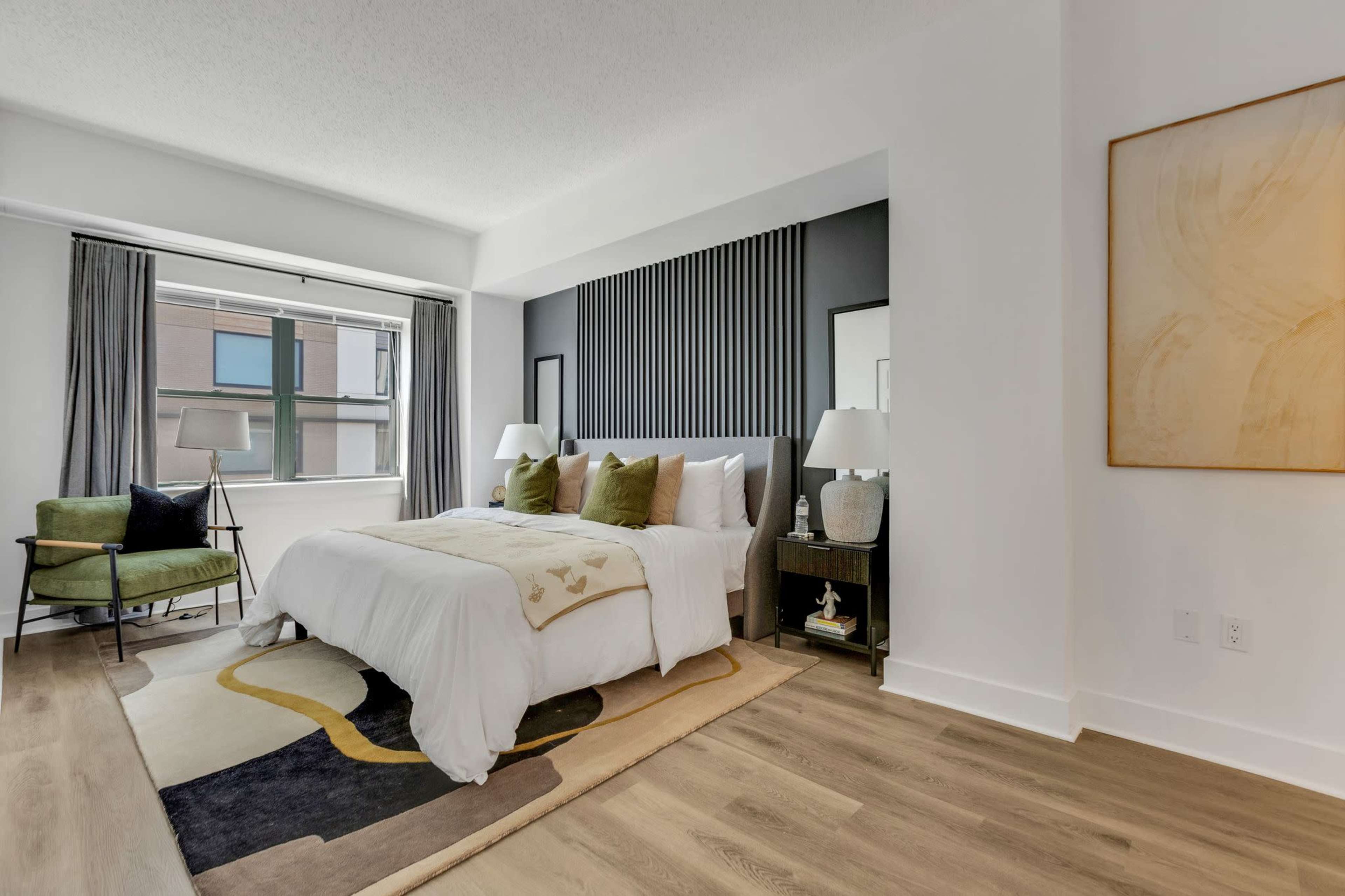 A modern bedroom with a bed, two bedside tables, a green armchair, and large windows letting in natural light.