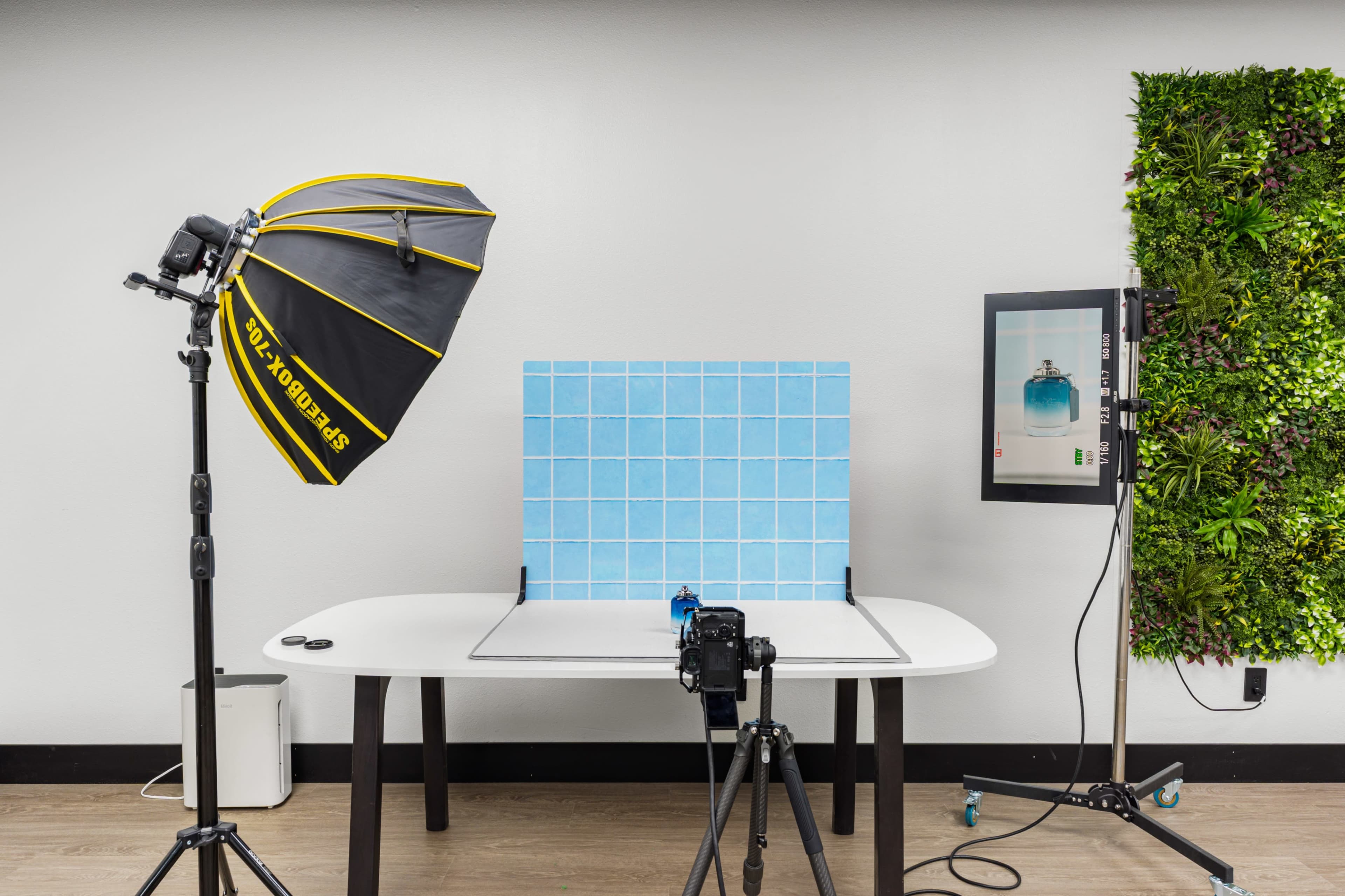 The image shows a photography setup with a blue tiled backdrop, a camera on a tripod, a softbox light, and a monitor displaying the subject, all arranged on a table in a studio environment.