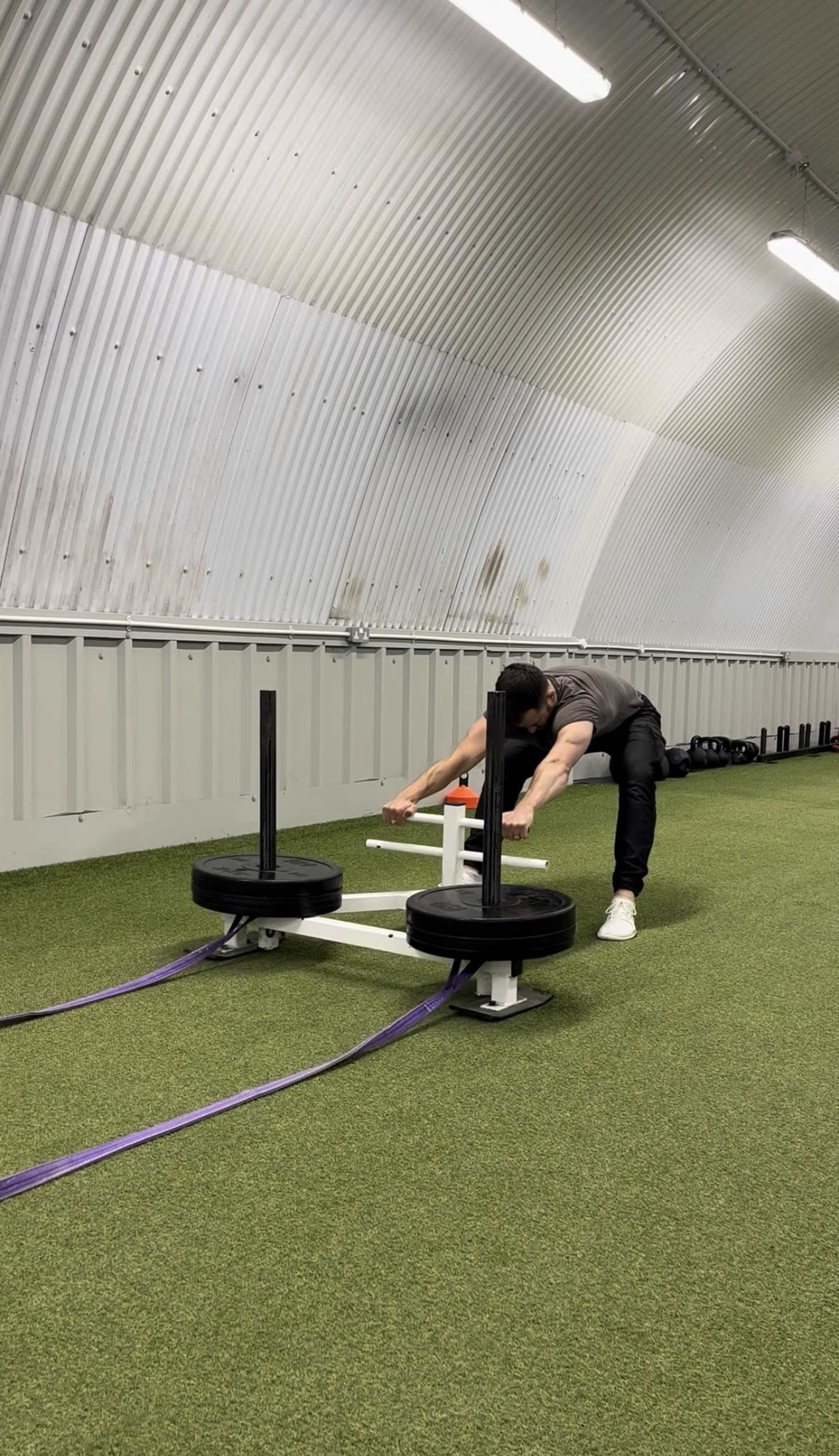 A person is pushing a sled with weights across a turf surface in a training facility.