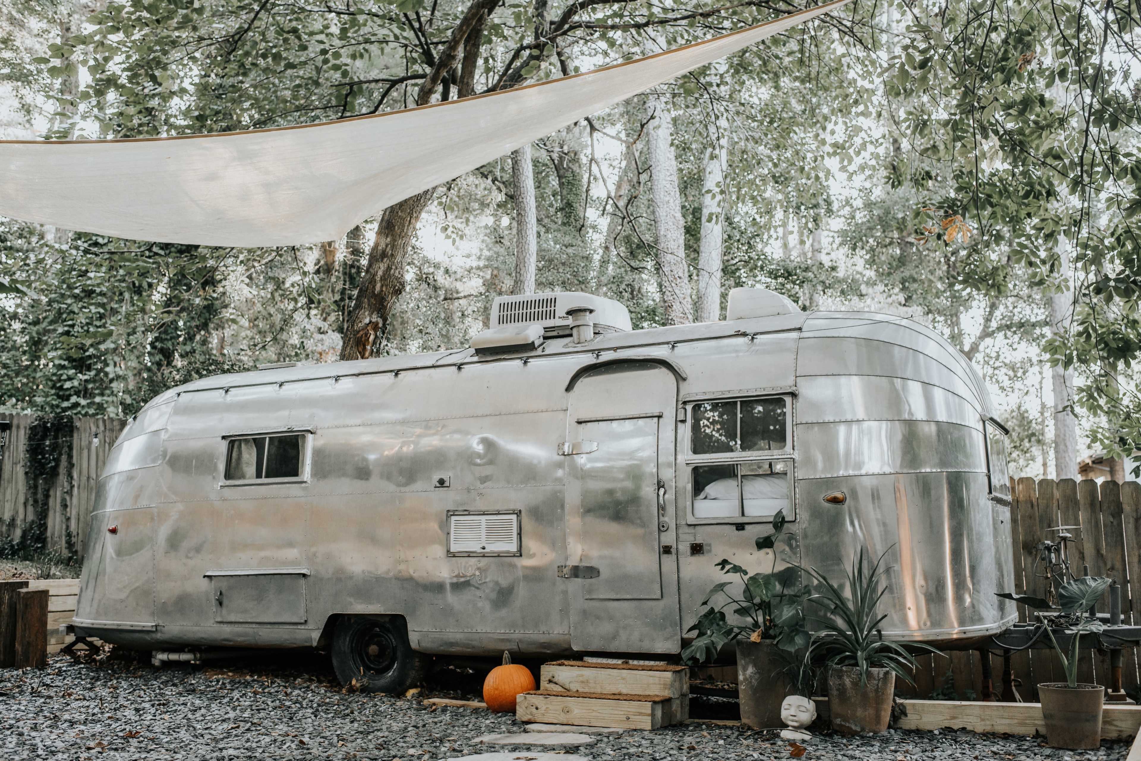 A silver Airstream trailer is parked in a shaded outdoor area surrounded by trees and gravel, with a few potted plants and a pumpkin nearby.