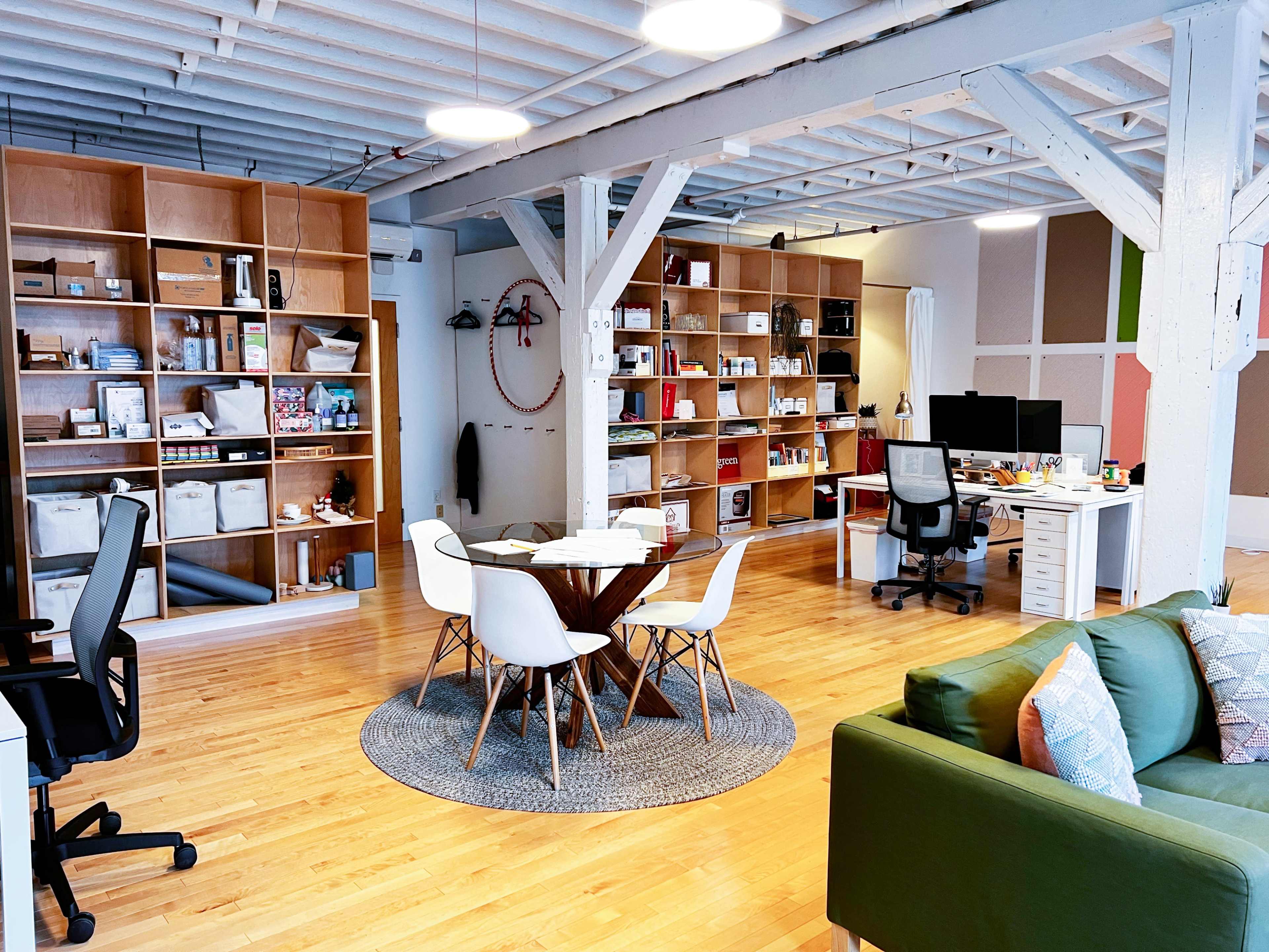 A modern open-plan office space with wooden floors, a round table surrounded by chairs, and shelves filled with various office supplies and decorative items.