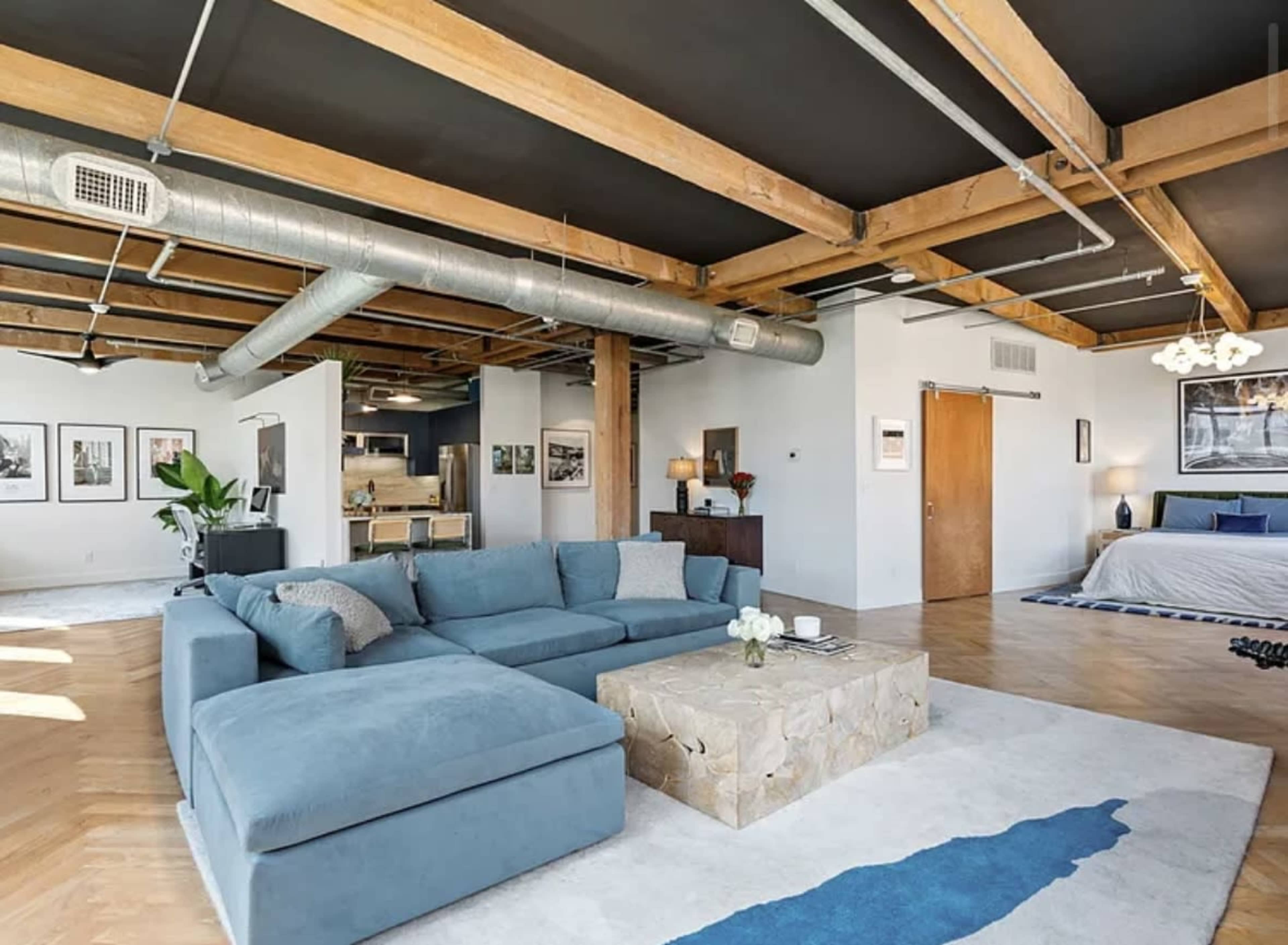 The image shows a spacious, modern living area featuring a large blue sectional sofa, a stone coffee table, and exposed wooden beams on the ceiling.