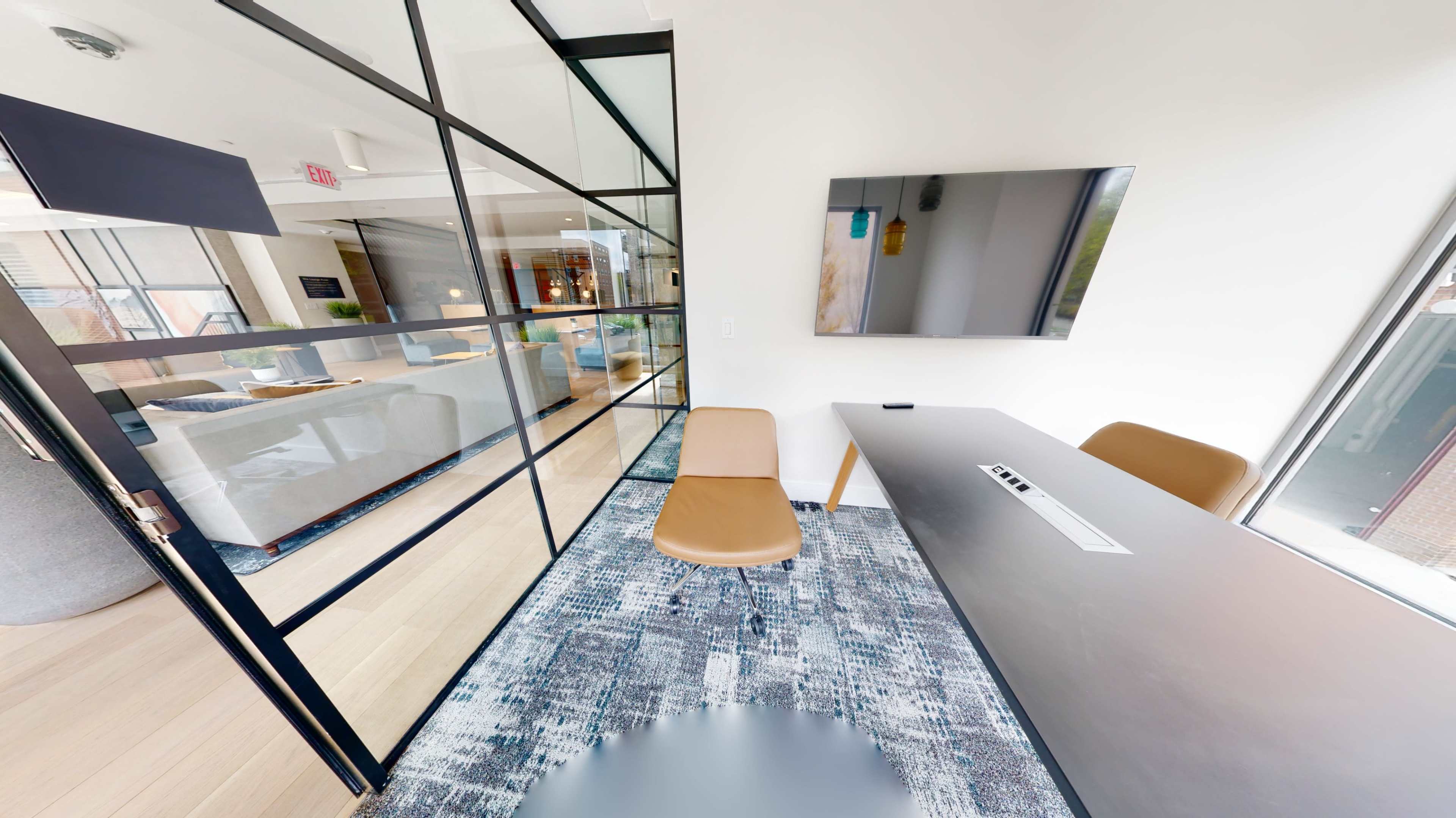 The image shows a modern office space featuring a glass-enclosed meeting room, a sleek desk, a chair, and a wall-mounted television.