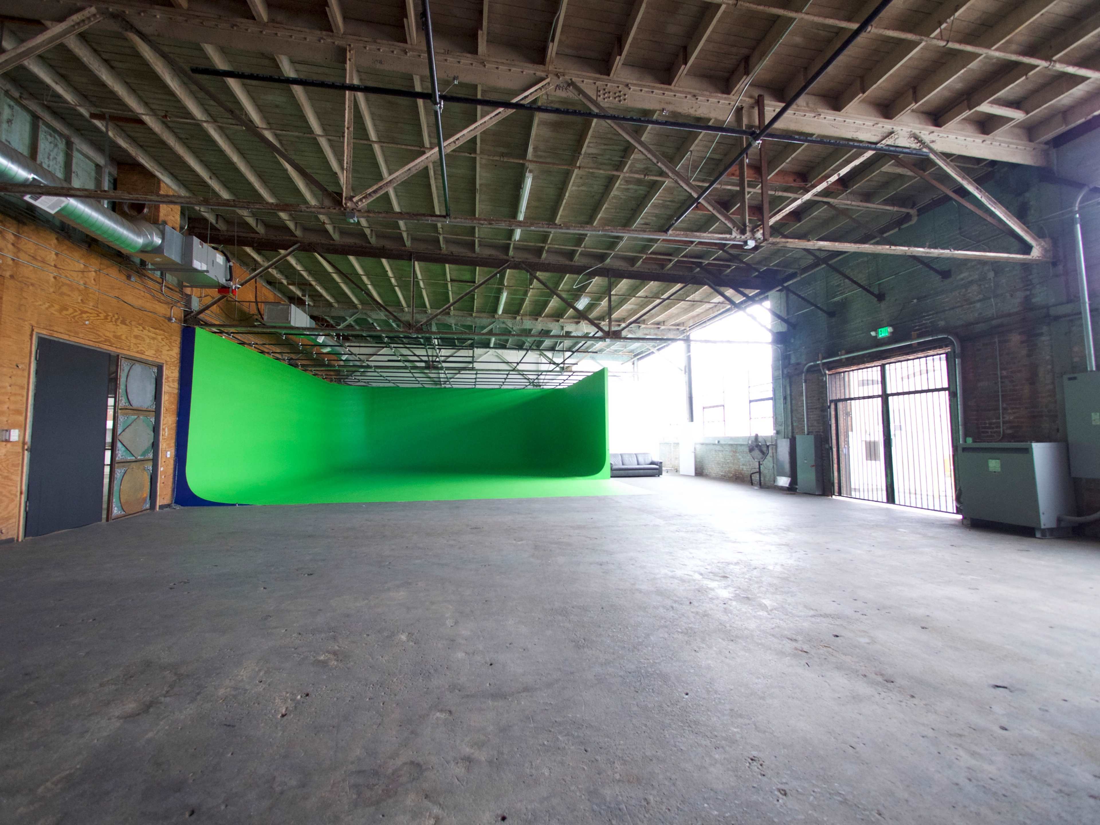 The interior of a spacious warehouse features a green screen backdrop against a wooden wall, with large windows allowing natural light to enter.