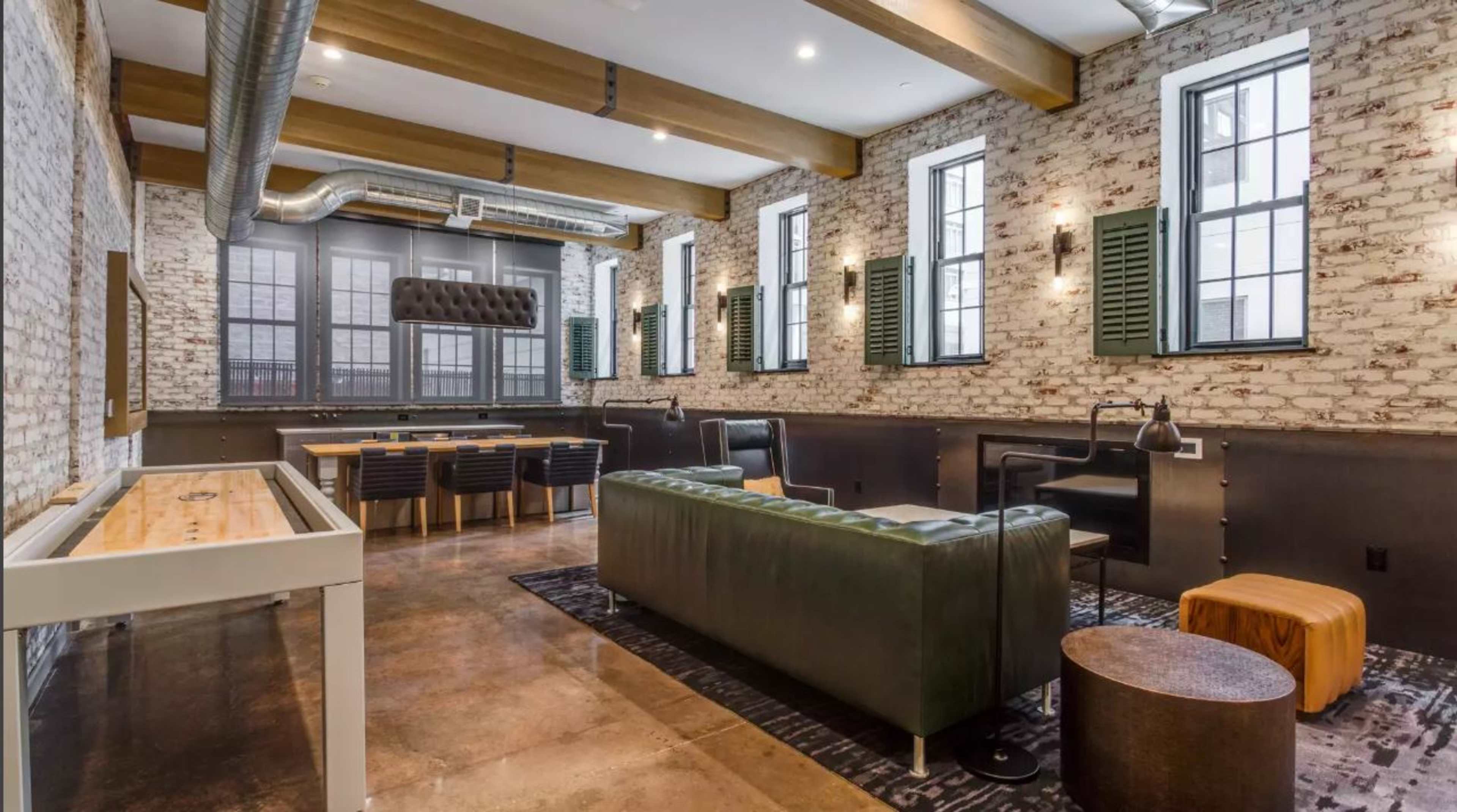 A spacious, industrial-style room with exposed brick walls, large windows, and a mix of seating arrangements including a green leather sofa and a wooden dining table.