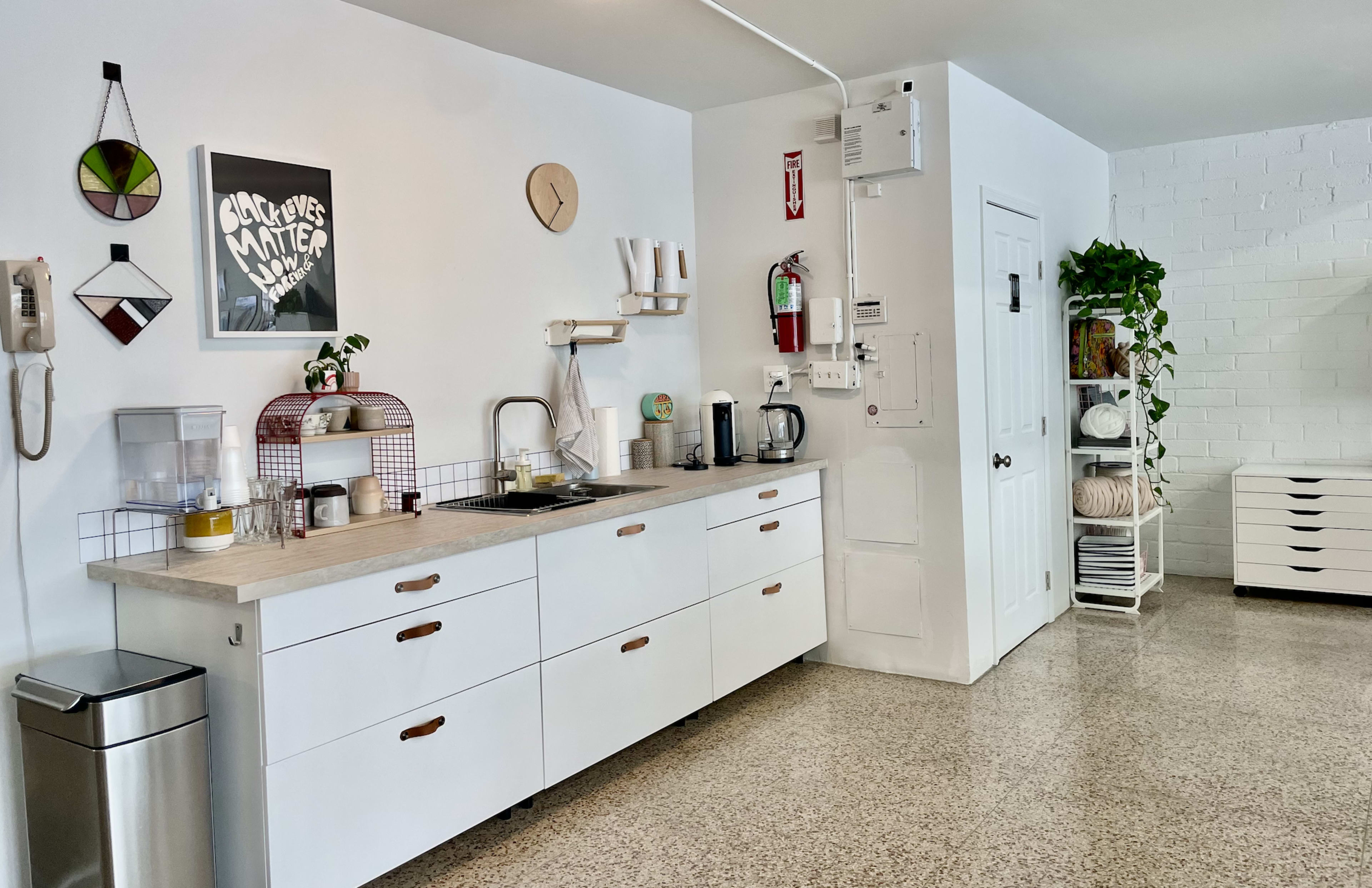 A modern kitchen space featuring white cabinetry, a sink, a countertop with kitchen appliances, and a wall adorned with decorative elements and a clock.