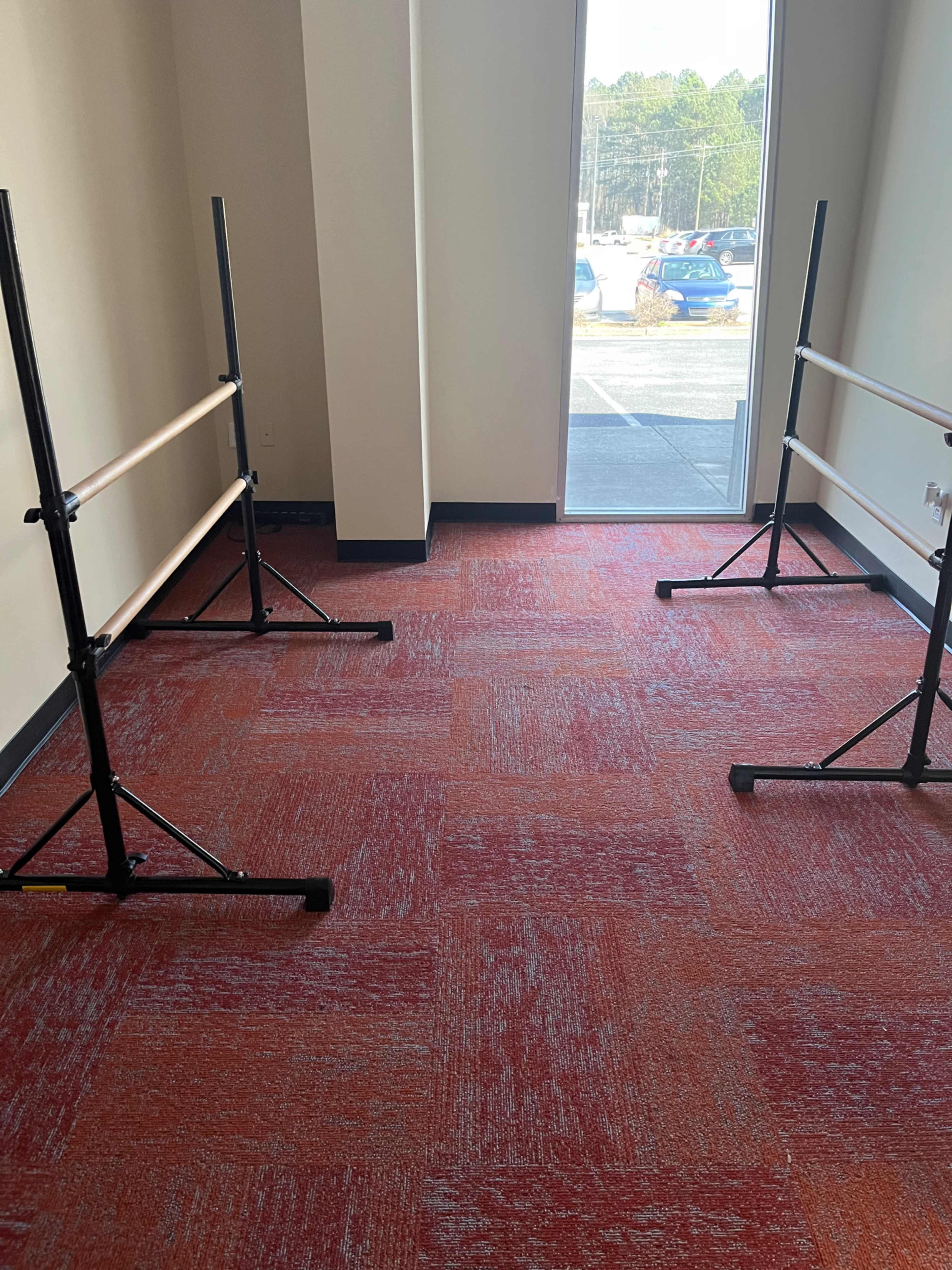 The image shows two ballet barres positioned parallel to each other in an empty room with red carpet flooring and a doorway leading outside.