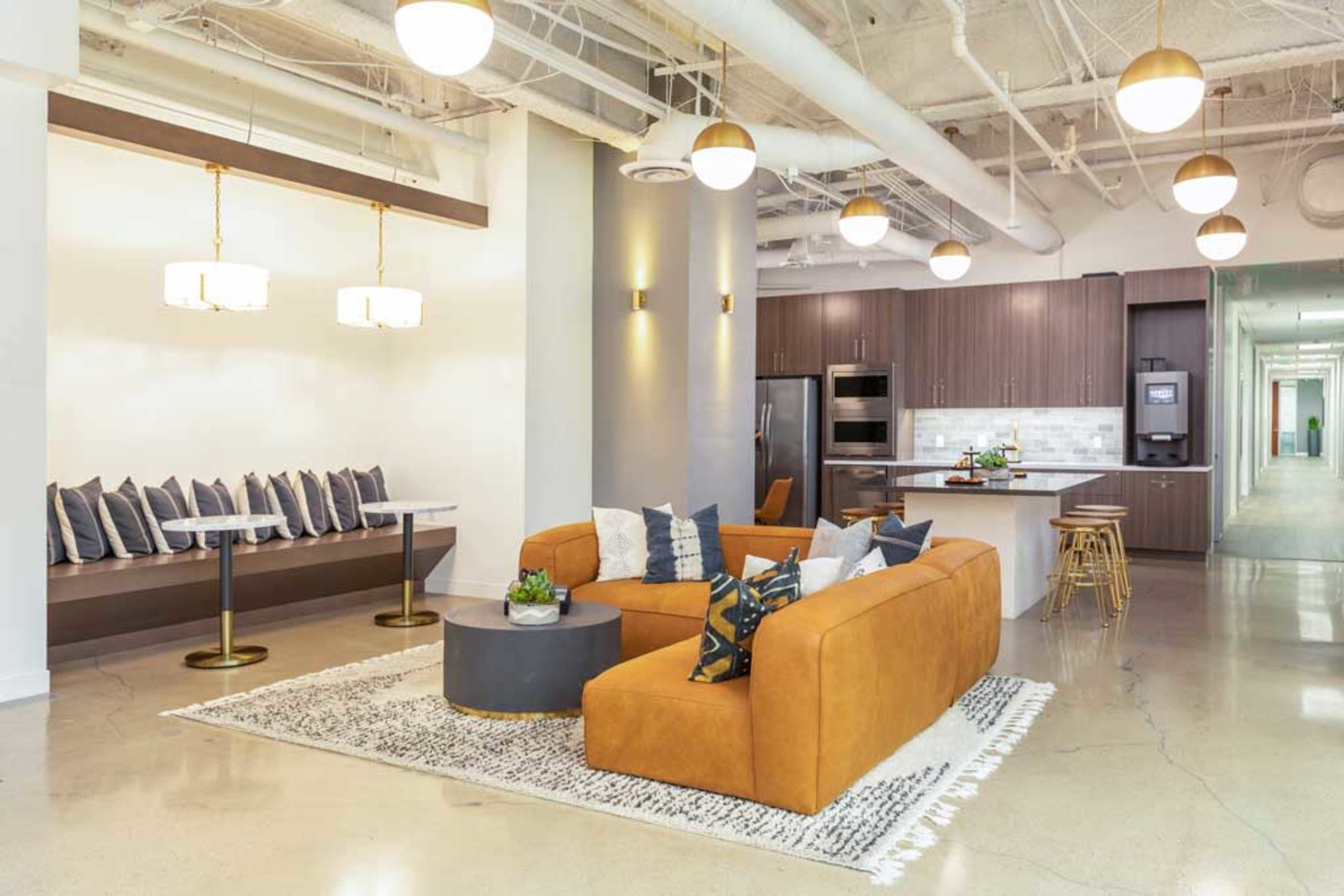 The image shows a modern lounge area with a large brown sectional sofa, decorative pillows, a round coffee table, and a kitchen in the background.